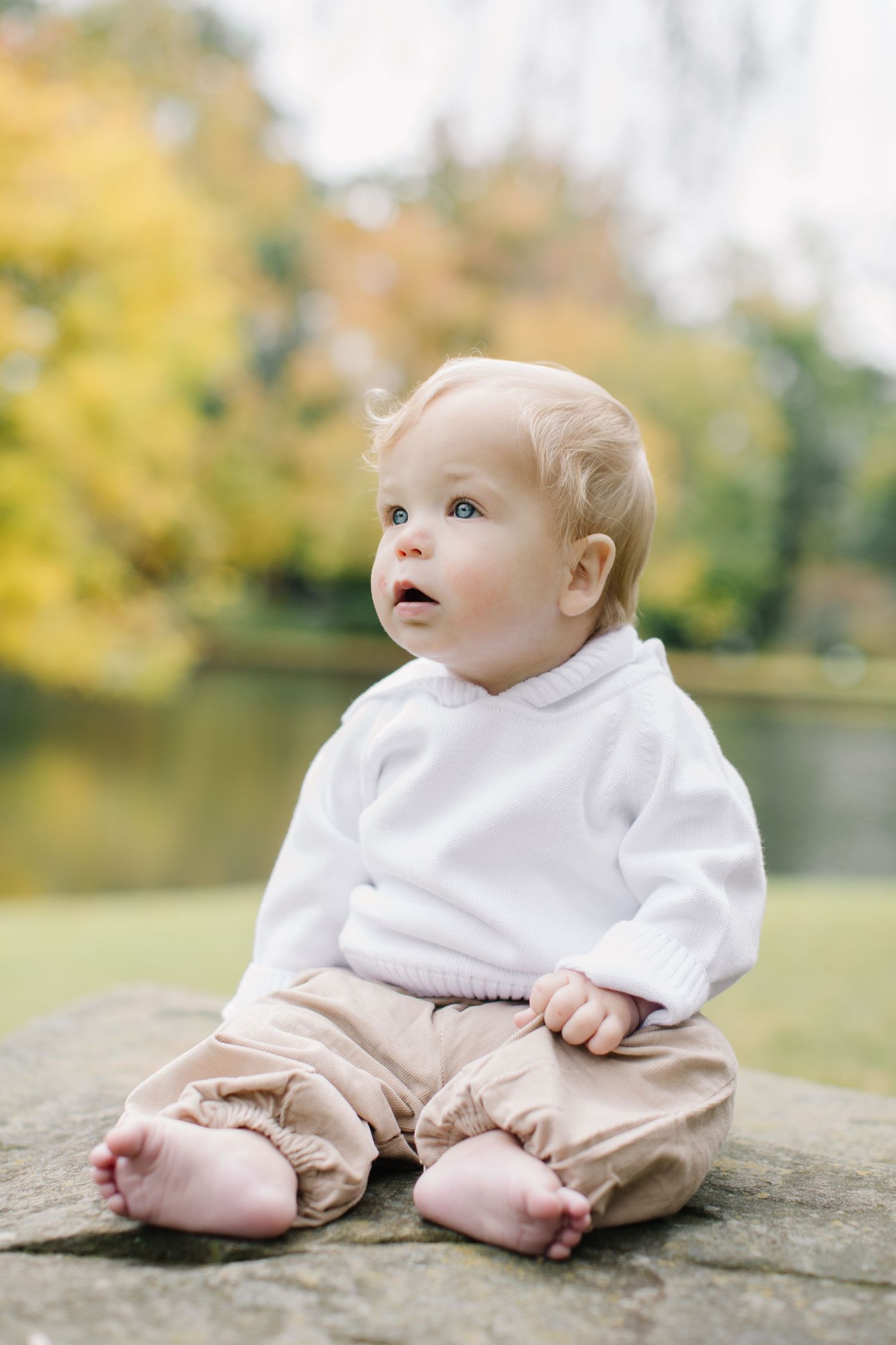 Baby in white sweater and beige pants sits on stone surface near pond with autumn foliage in background.