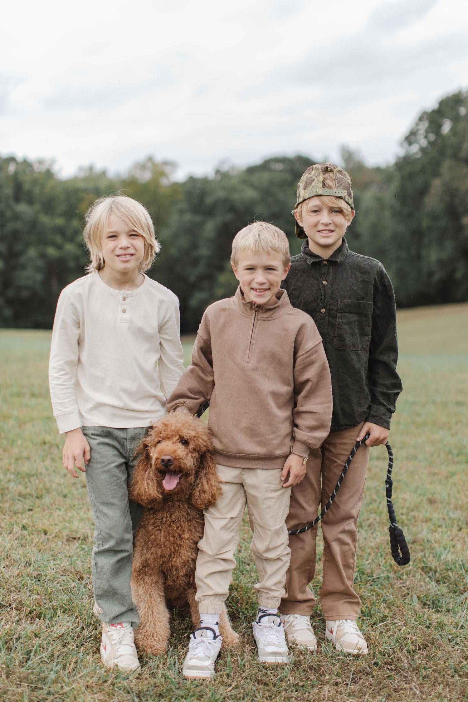 Three children in neutral colored clothing stand together with their goldendoodle dog in a grassy field.