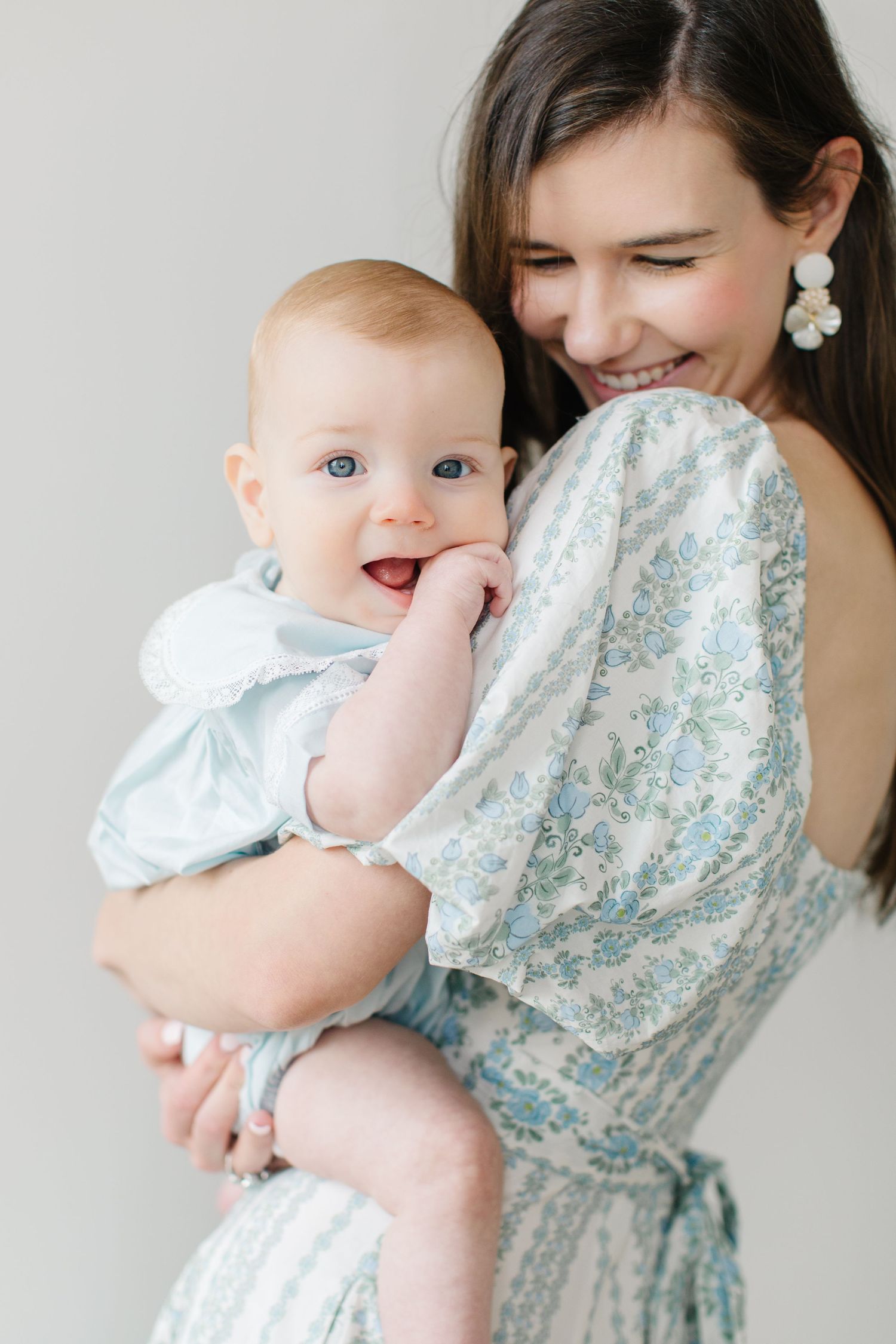 A smiling mother in a floral dress holds her happy baby in a light blue outfit against a soft white background.
