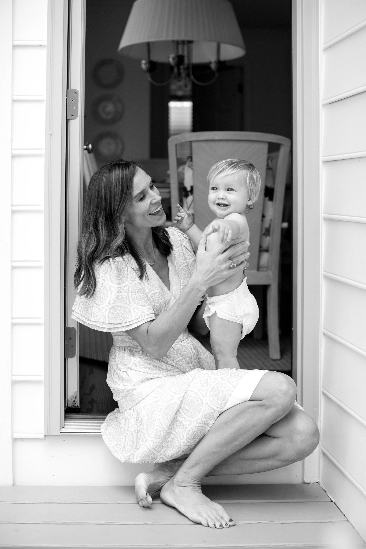 Loving moment between a mother and baby in white dresses sitting by a window in black and white portraits.