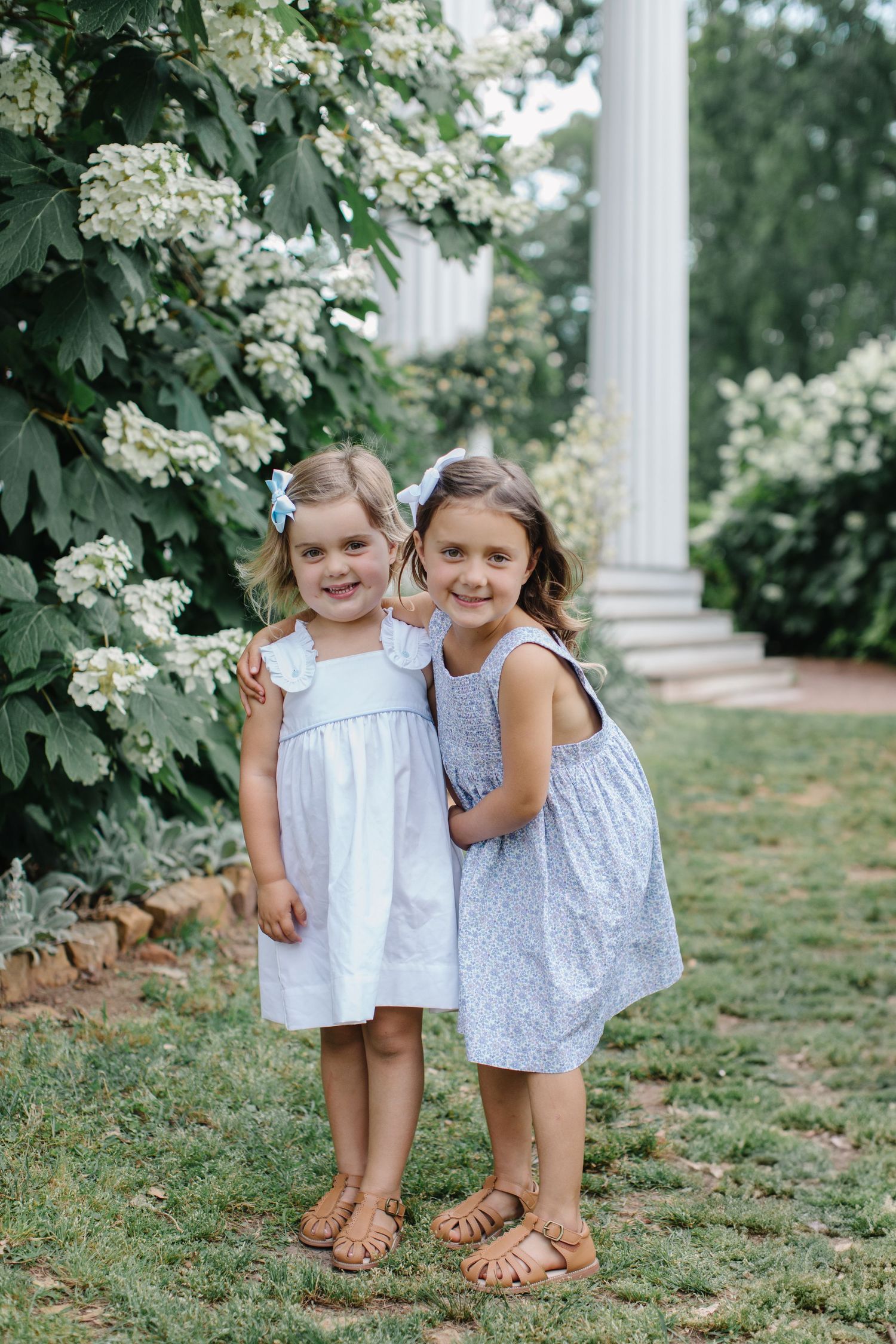 Two young girls in white and blue dresses pose together on grass near white flowering bushes and classic columns.