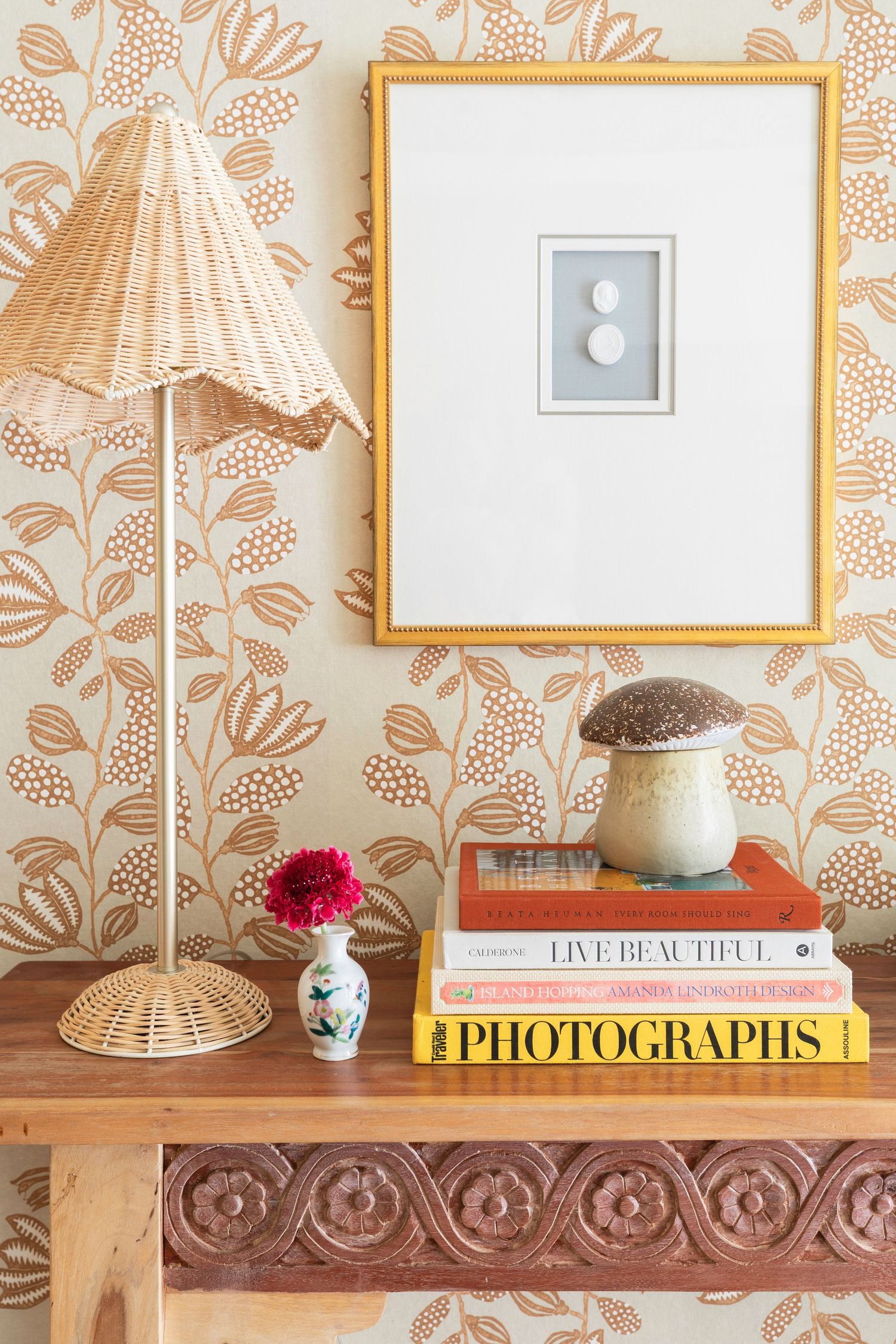 Boho console table with rattan lamp, stacked books, mushroom figurine, and floral wallpaper backdrop in a stylish home decor setup.