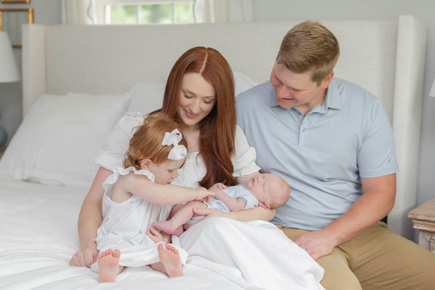 Family sitting together on white bed in light-filled bedroom with parents holding two small children.