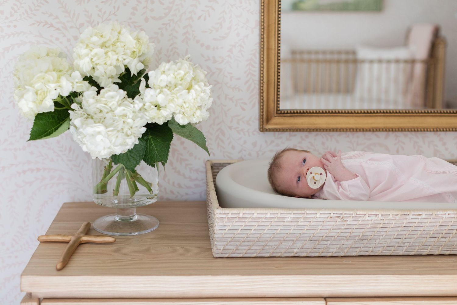 White hydrangea flowers arranged in a clear glass vase sit elegantly on a wooden surface near framed artwork.