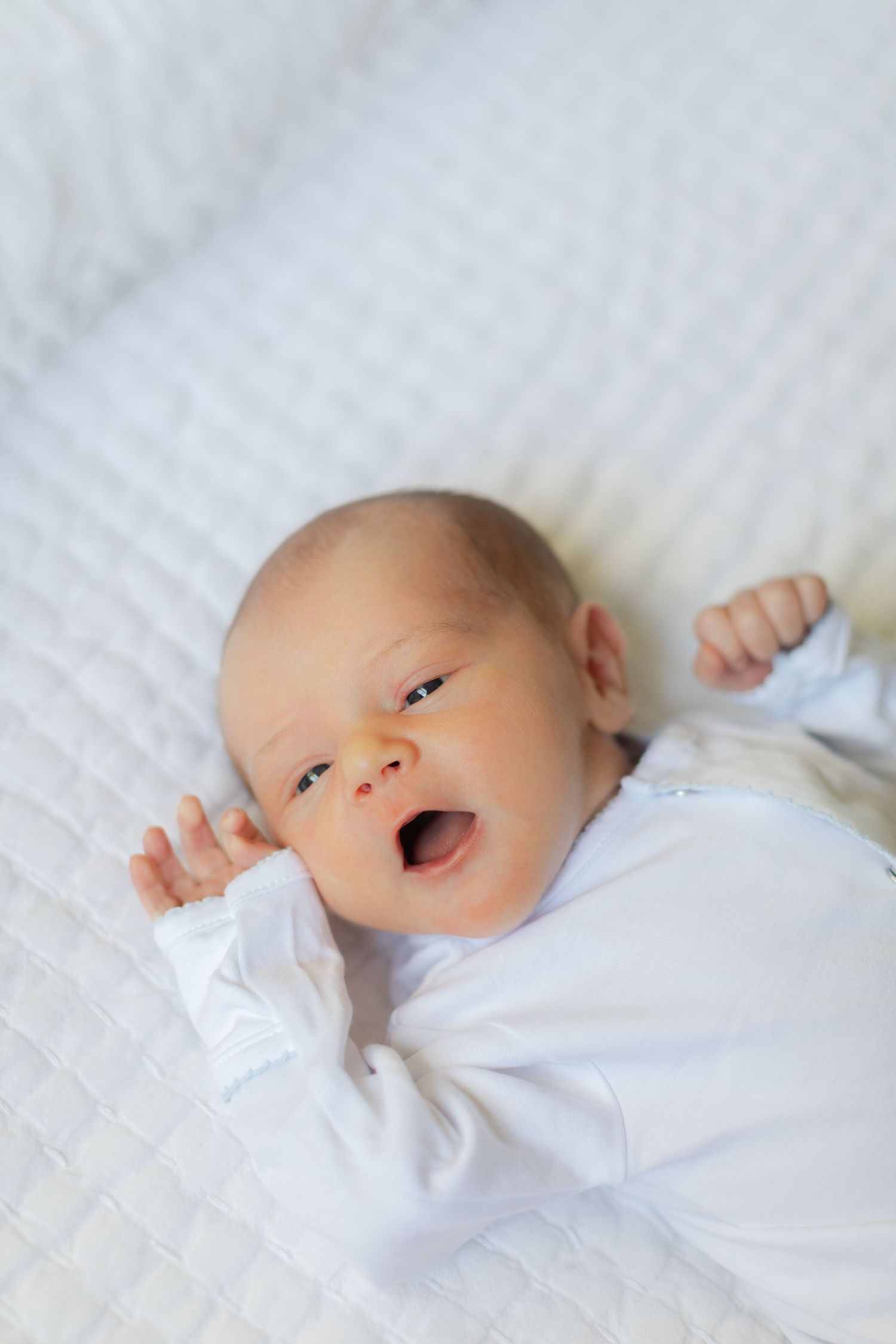 Newborn baby in white onesie lying on white blanket with mouth open and arms raised.