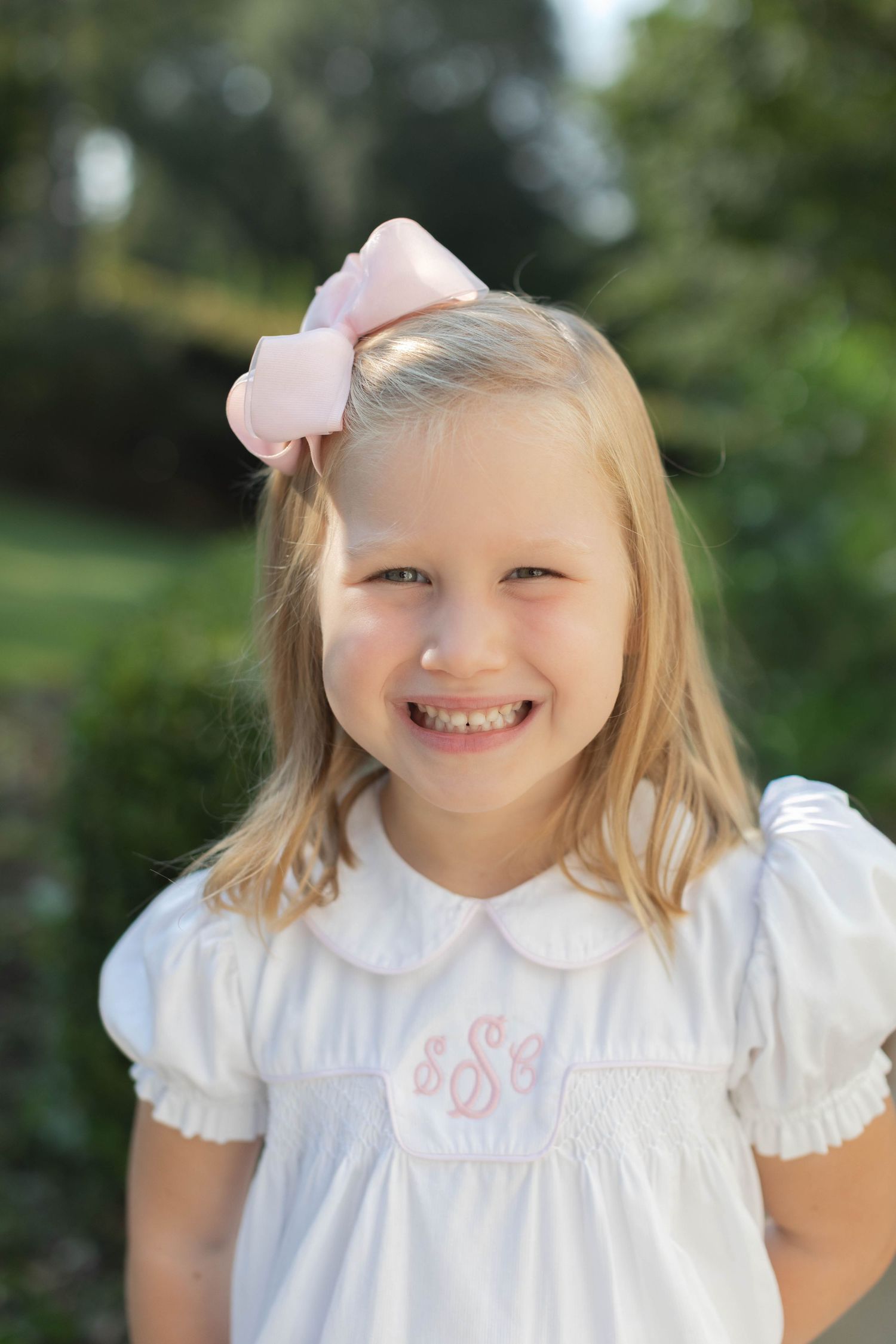 A young child wearing a white peter pan collar dress and pink hair bow outdoors in a natural setting.