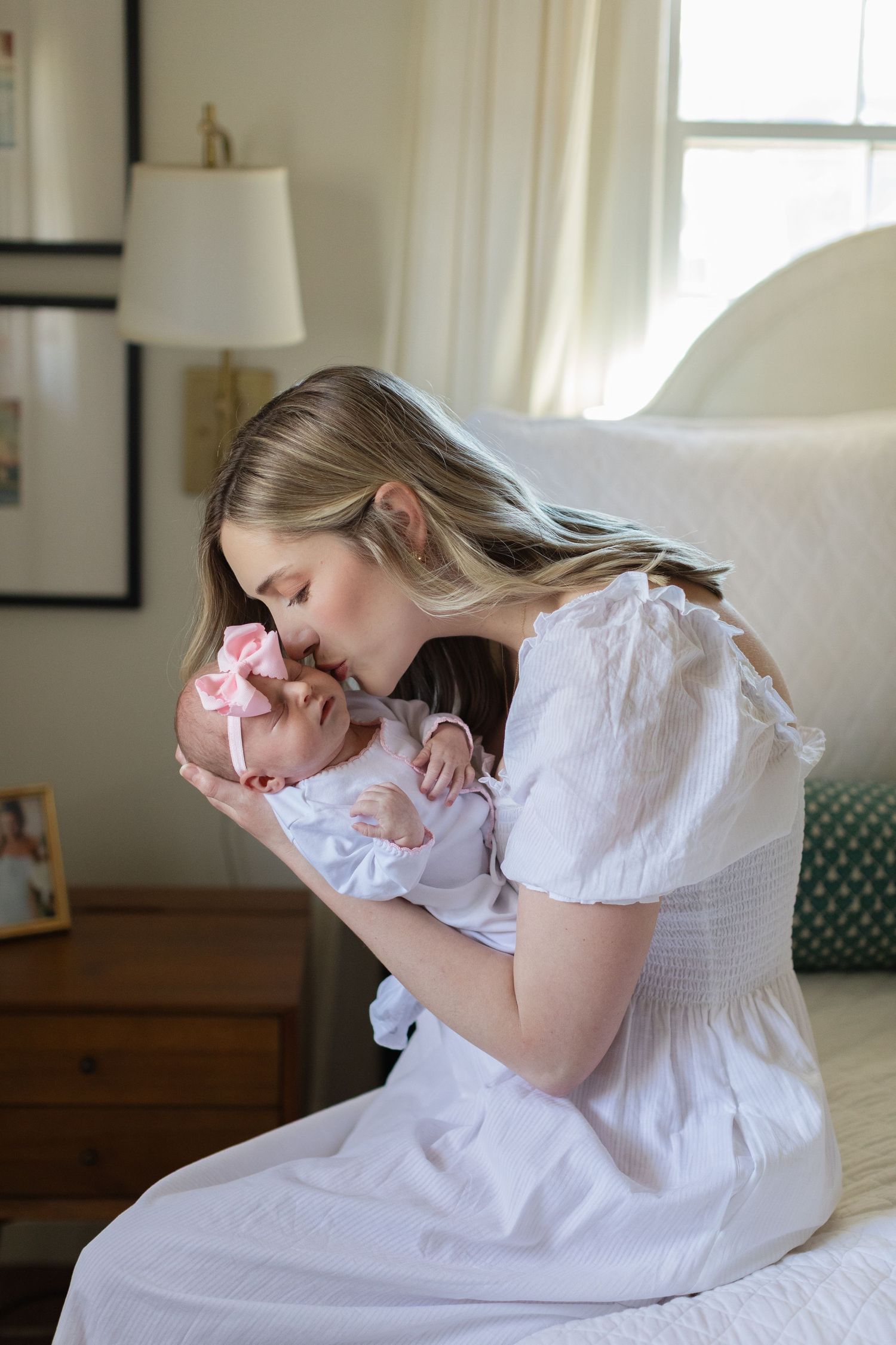 Woman in white dress cradles newborn on bed in sunlit room.