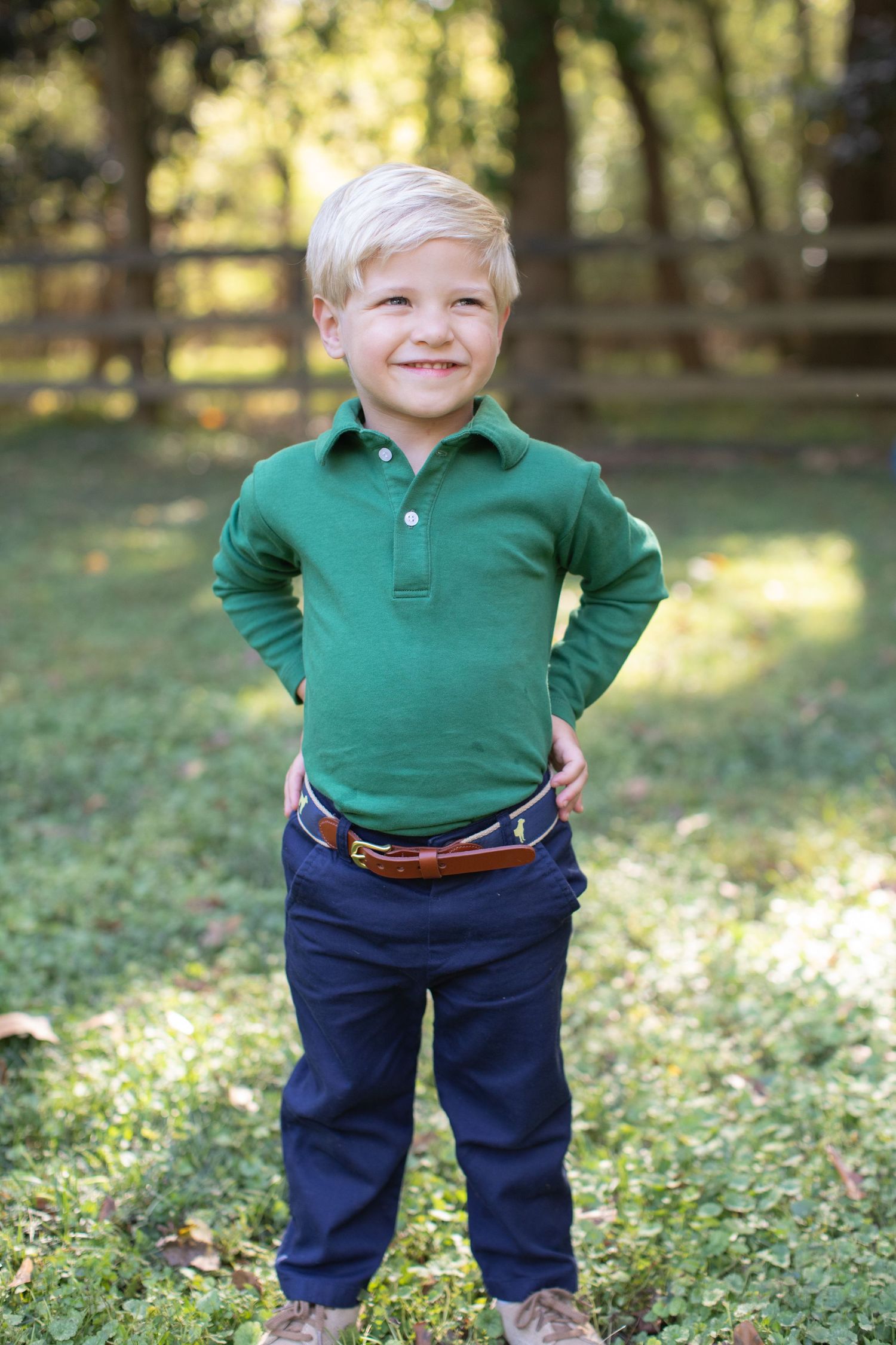 A small child wearing a green polo shirt and navy pants stands on grass in a sunlit outdoor setting.