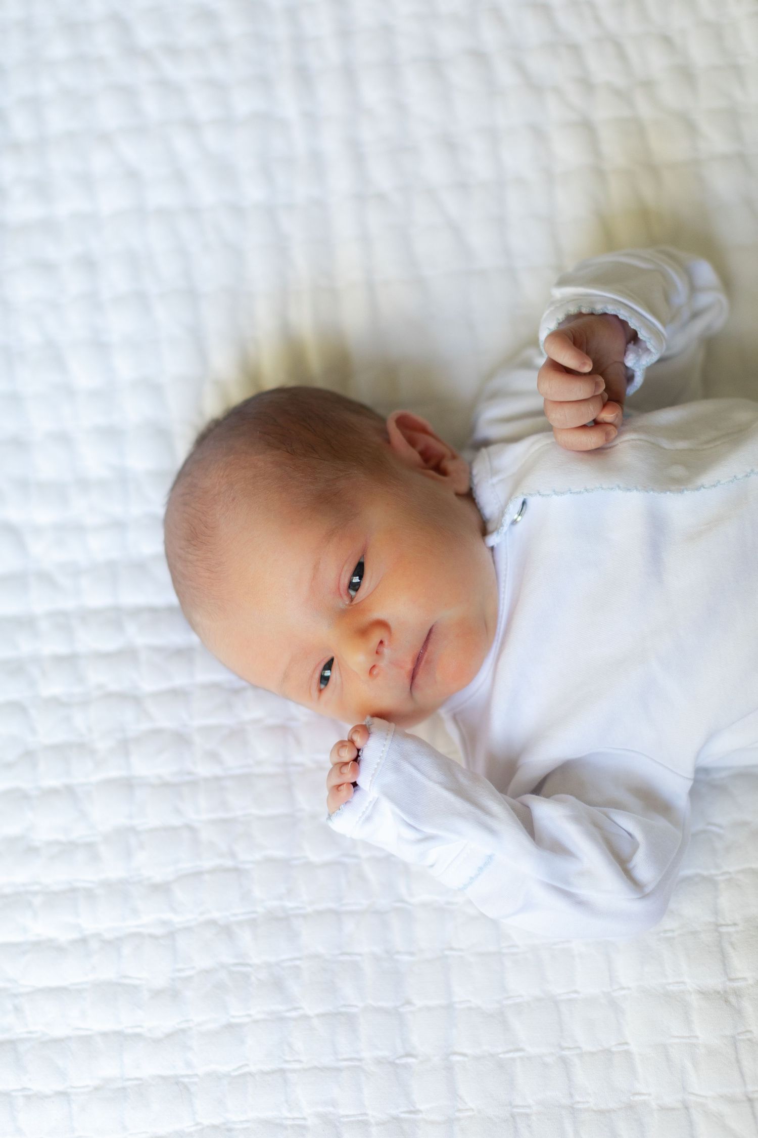 Peaceful newborn baby in white onesie lying on white quilted blanket with tiny fists raised, looking at camera.
