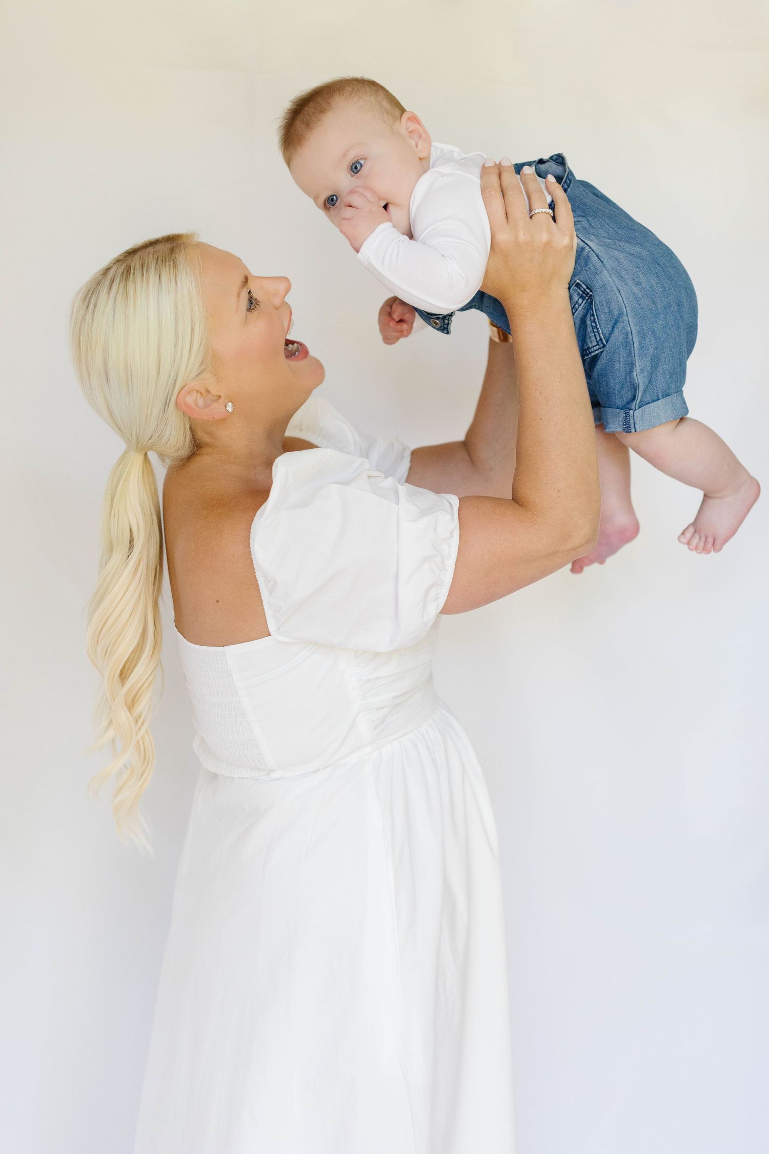 Parent in white dress lifts and plays with baby wearing white onesie against neutral background.