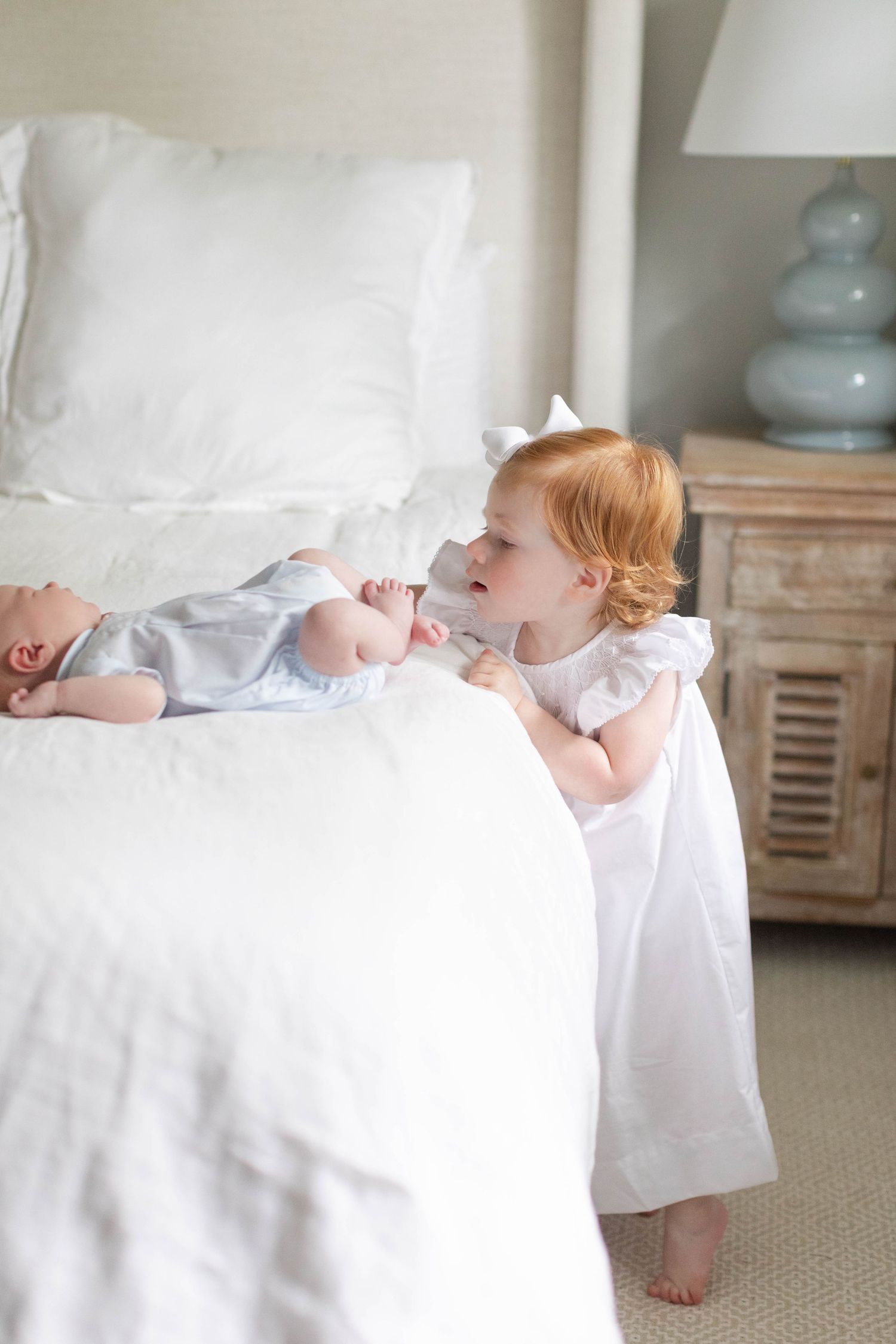 A small child in a white dress peers over the edge of a bed at something.