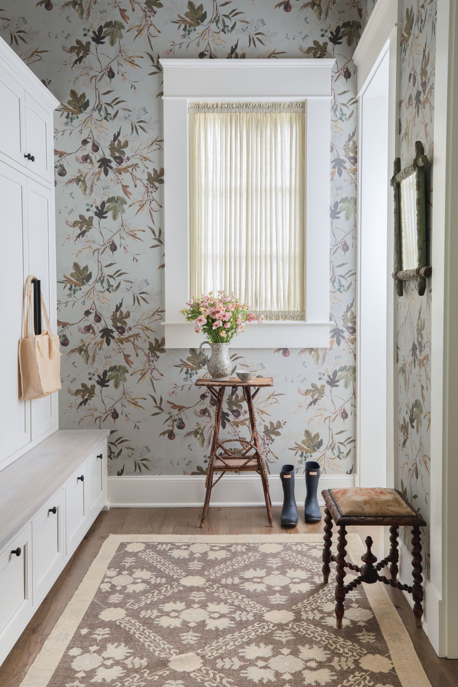 Elegant mudroom with botanical wallpaper, white built-ins, wooden stool with pink flowers, blue rain boots, and a patterned area rug.