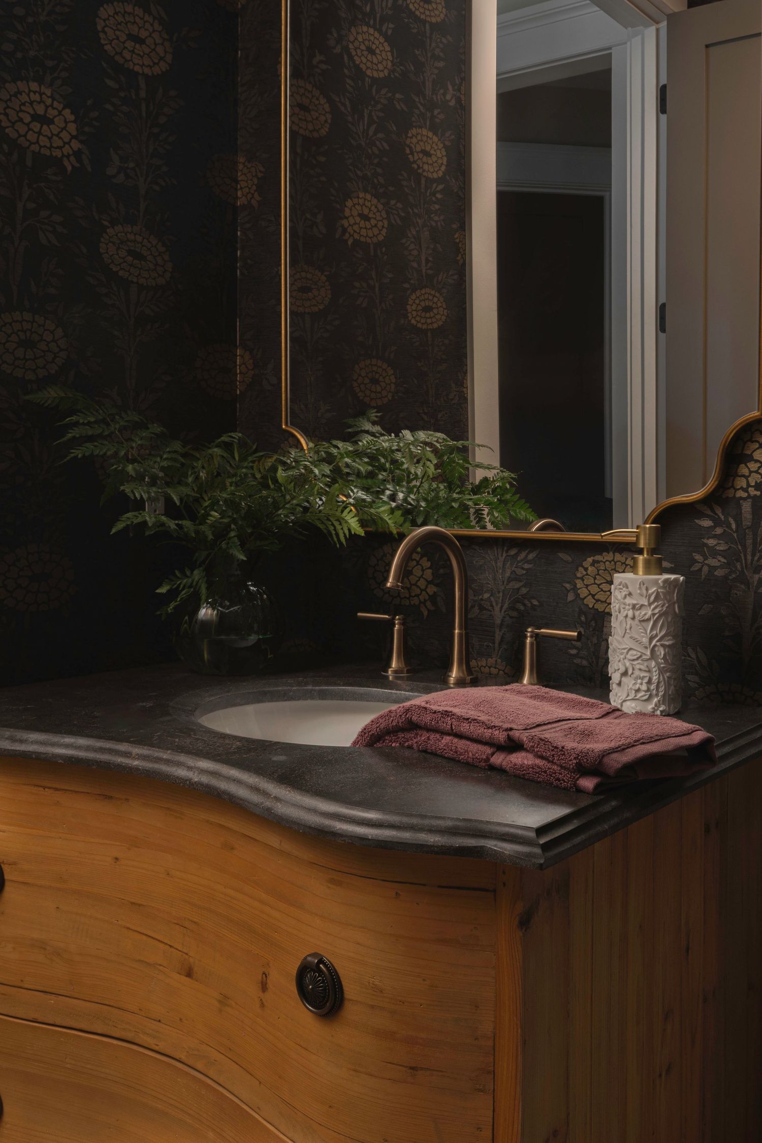 Vintage wooden bathroom vanity with copper faucet, stone countertop, burgundy towel and potted fern against floral wallpaper.