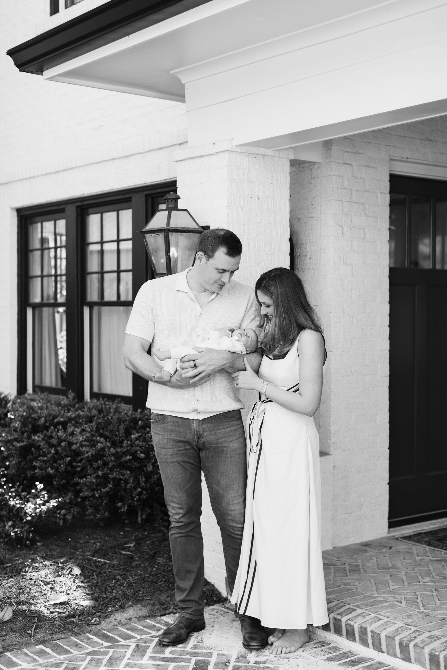 Black and white photo of couple admiring newborn baby outside elegant brick home with lantern.