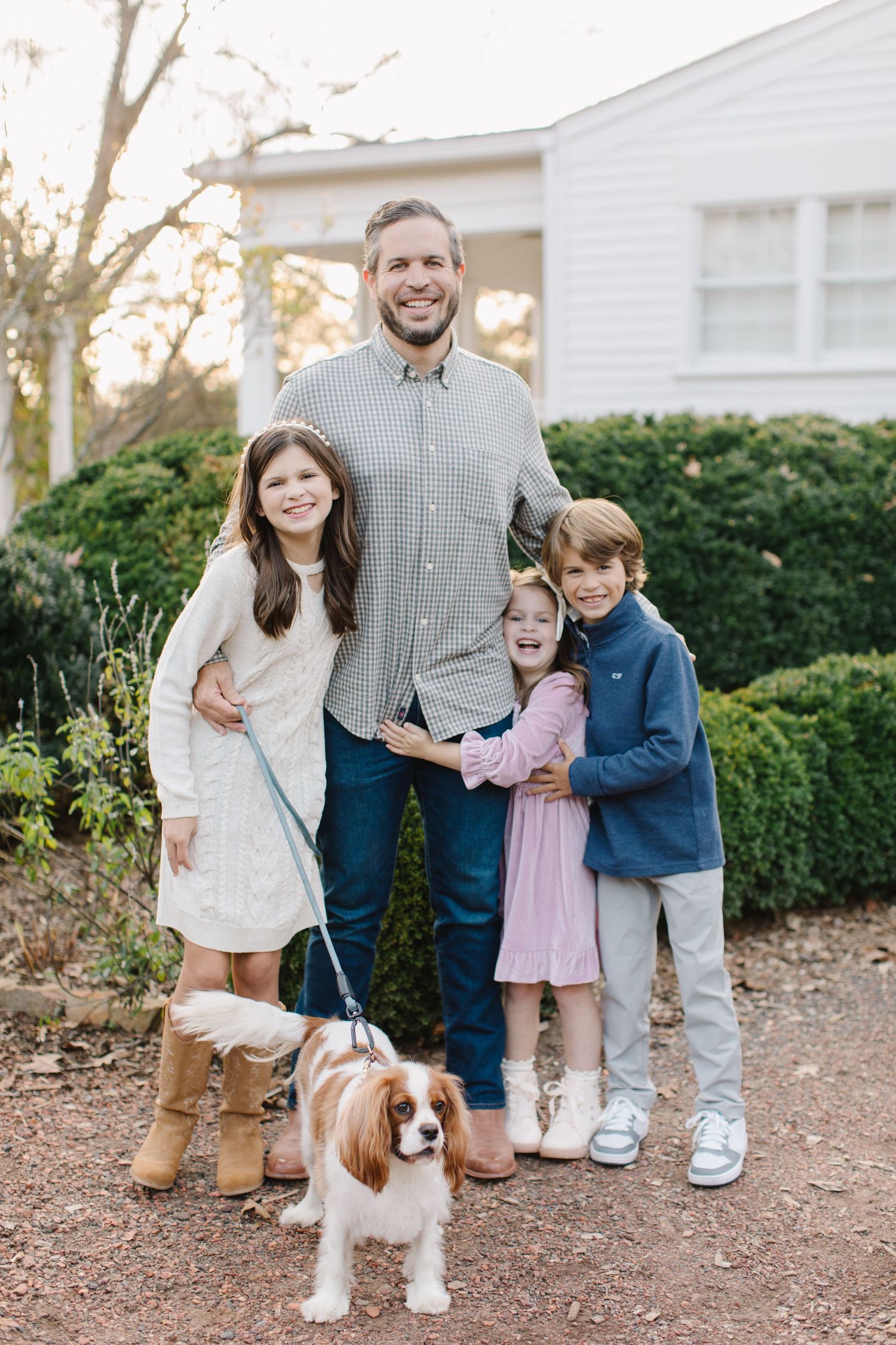 A family poses outdoors with their Cavalier King Charles Spaniel dog on a gravel path near green hedges.