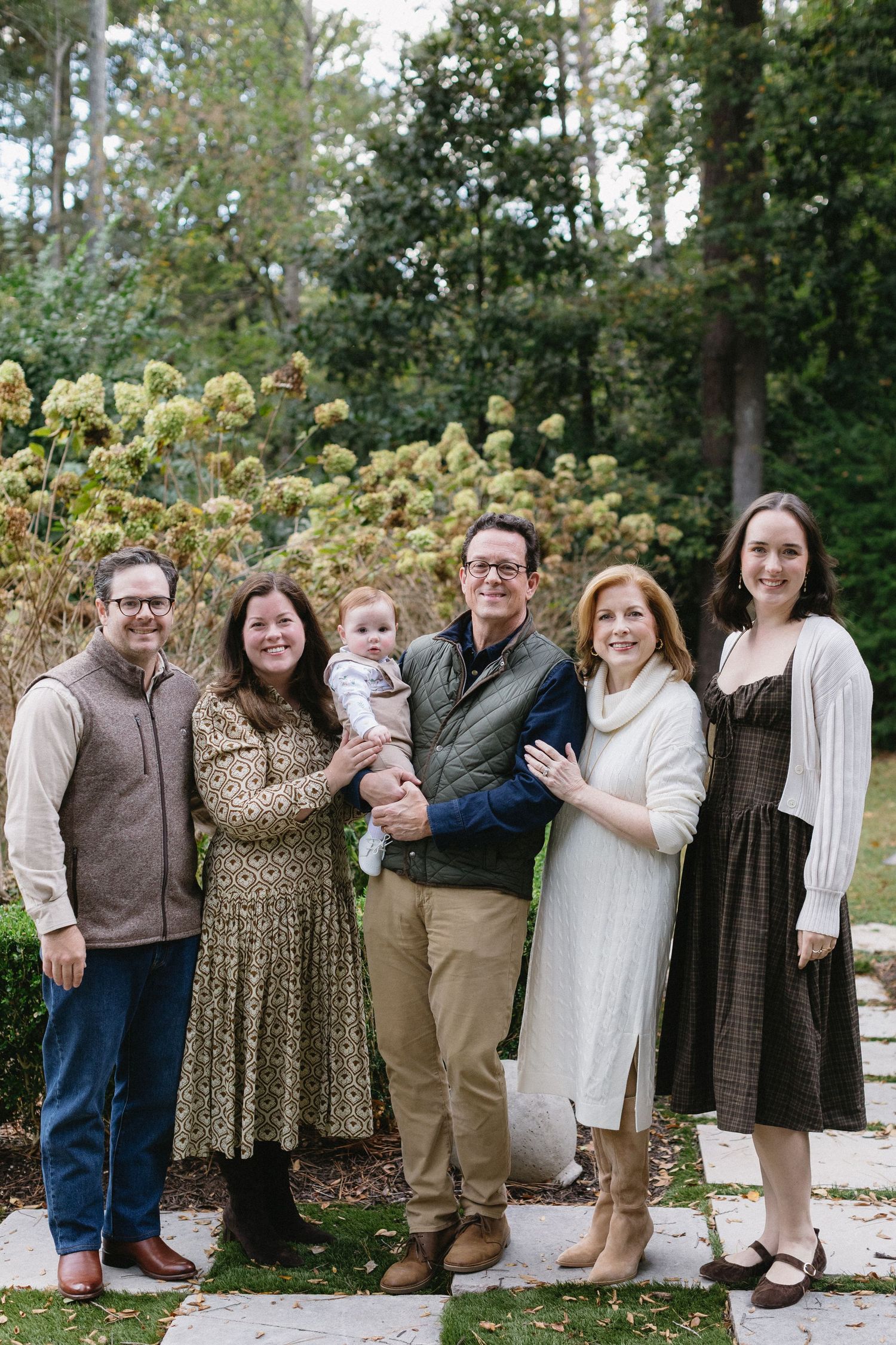 A group of people pose together outdoors against a backdrop of green trees and foliage in casual attire.