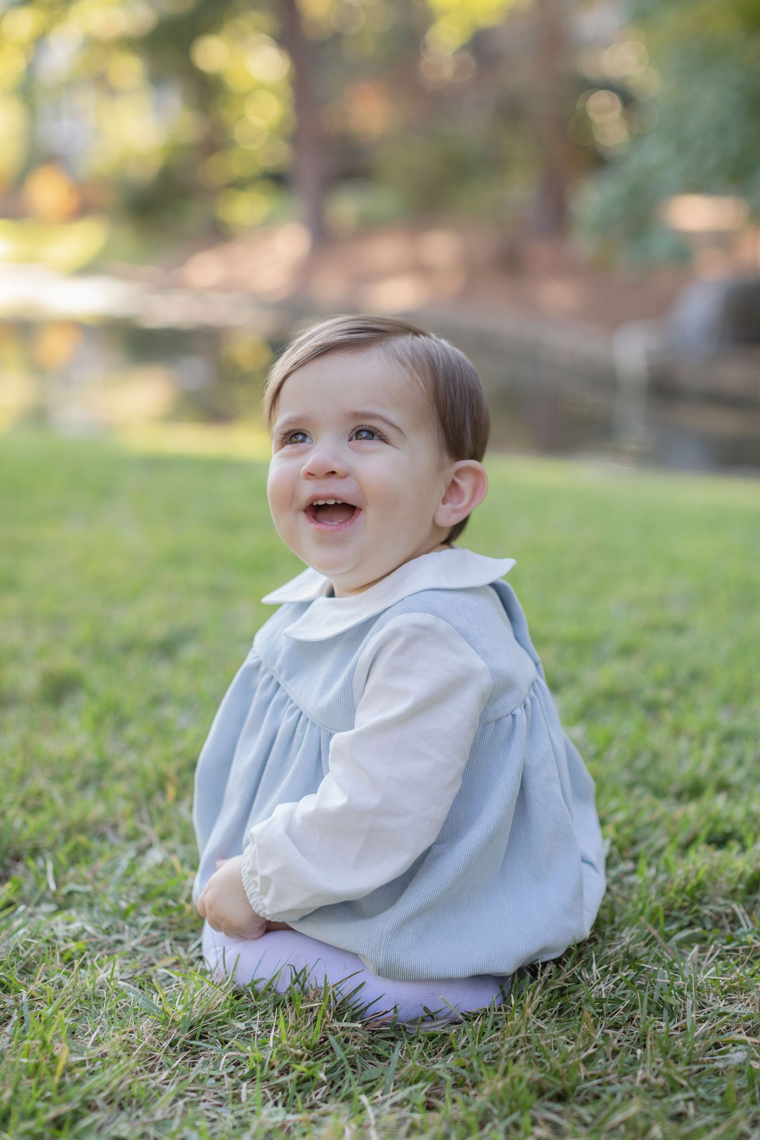 Baby in a white sweater sits on grass and looks up with a joyful smile on a sunny day.