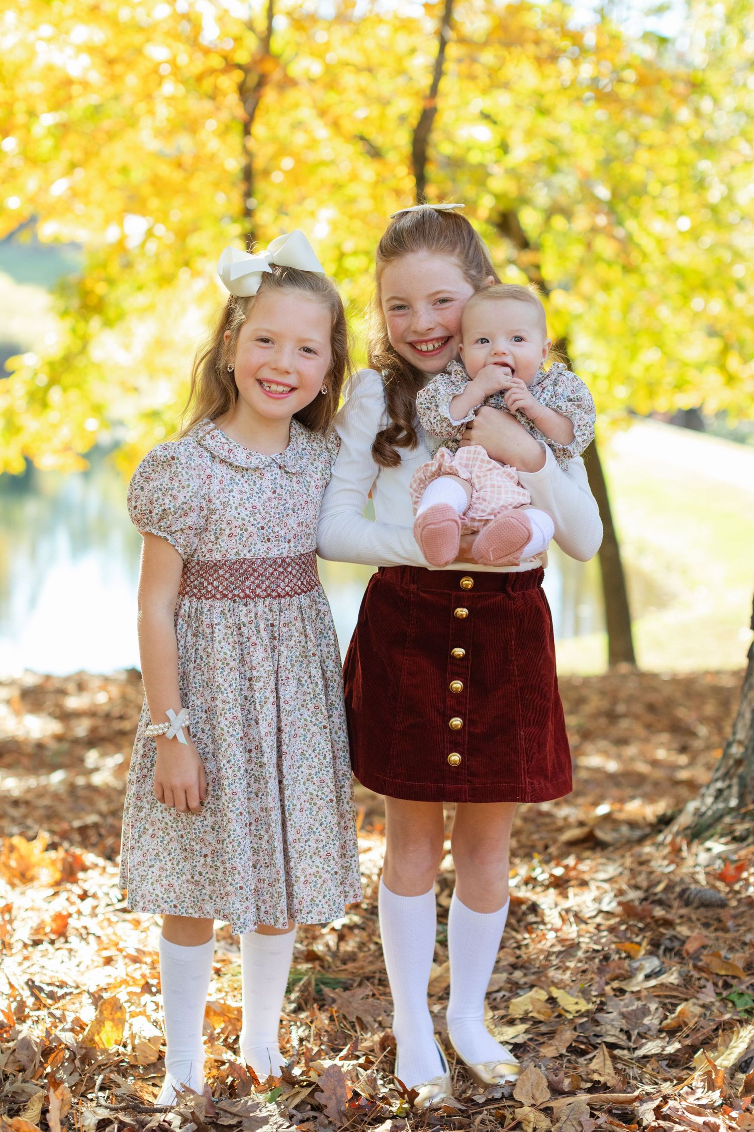 Three children pose together on fallen autumn leaves with bright yellow foliage in the background.