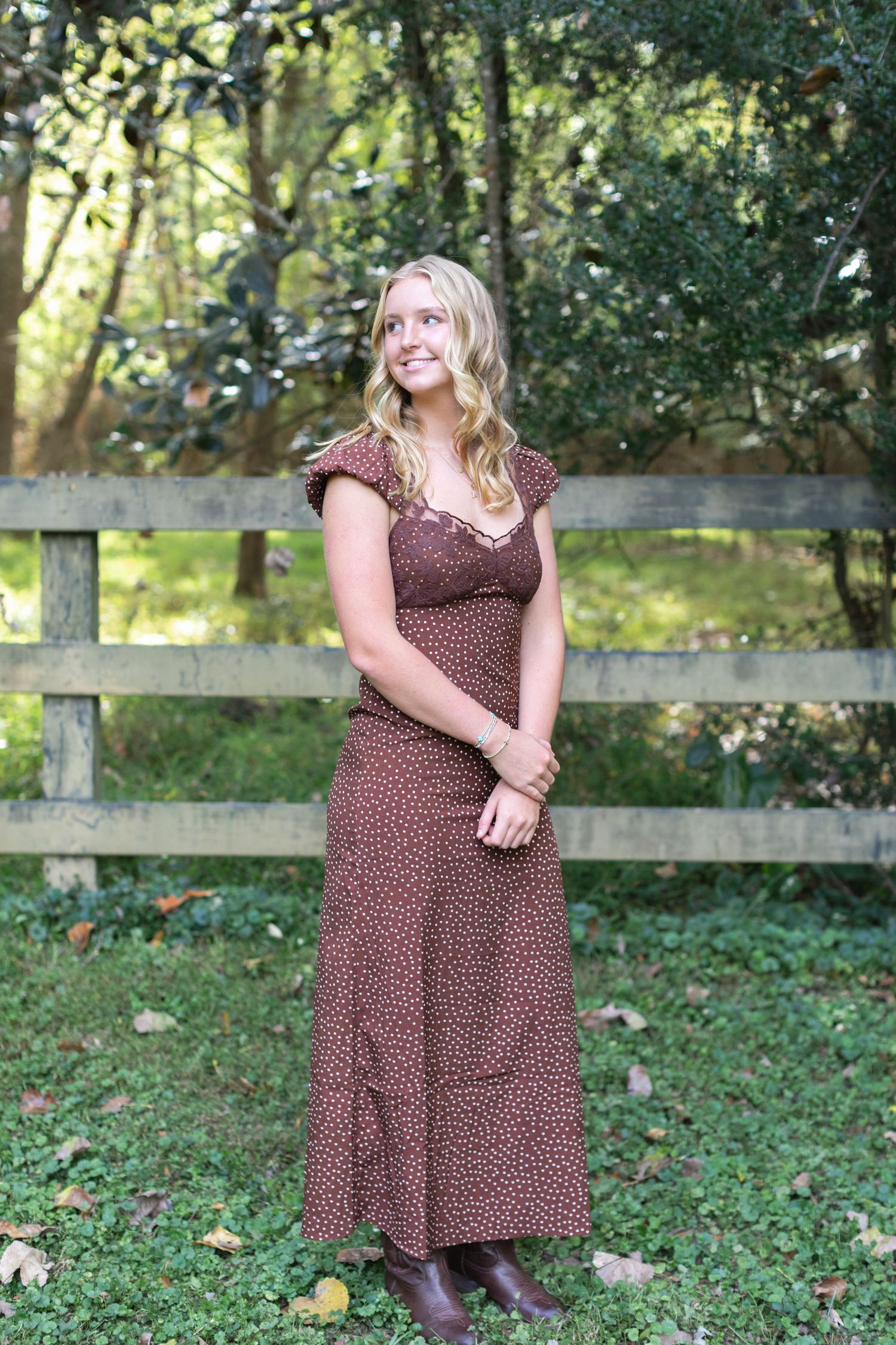 A person in a brown polka dot dress poses outdoors against a rustic wooden fence and green foliage.