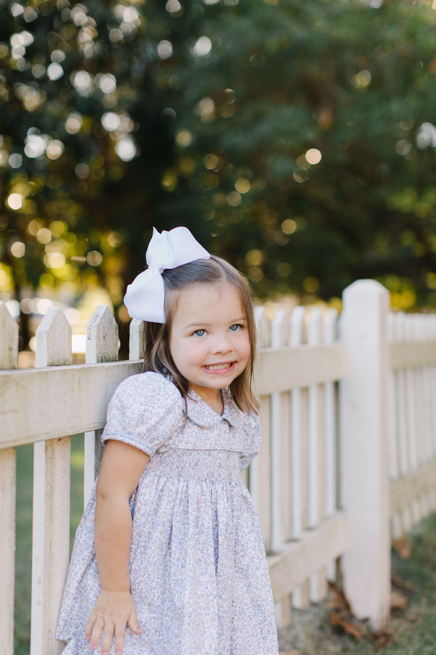 A small child in a white dress plays by a white picket fence on a sunny day.