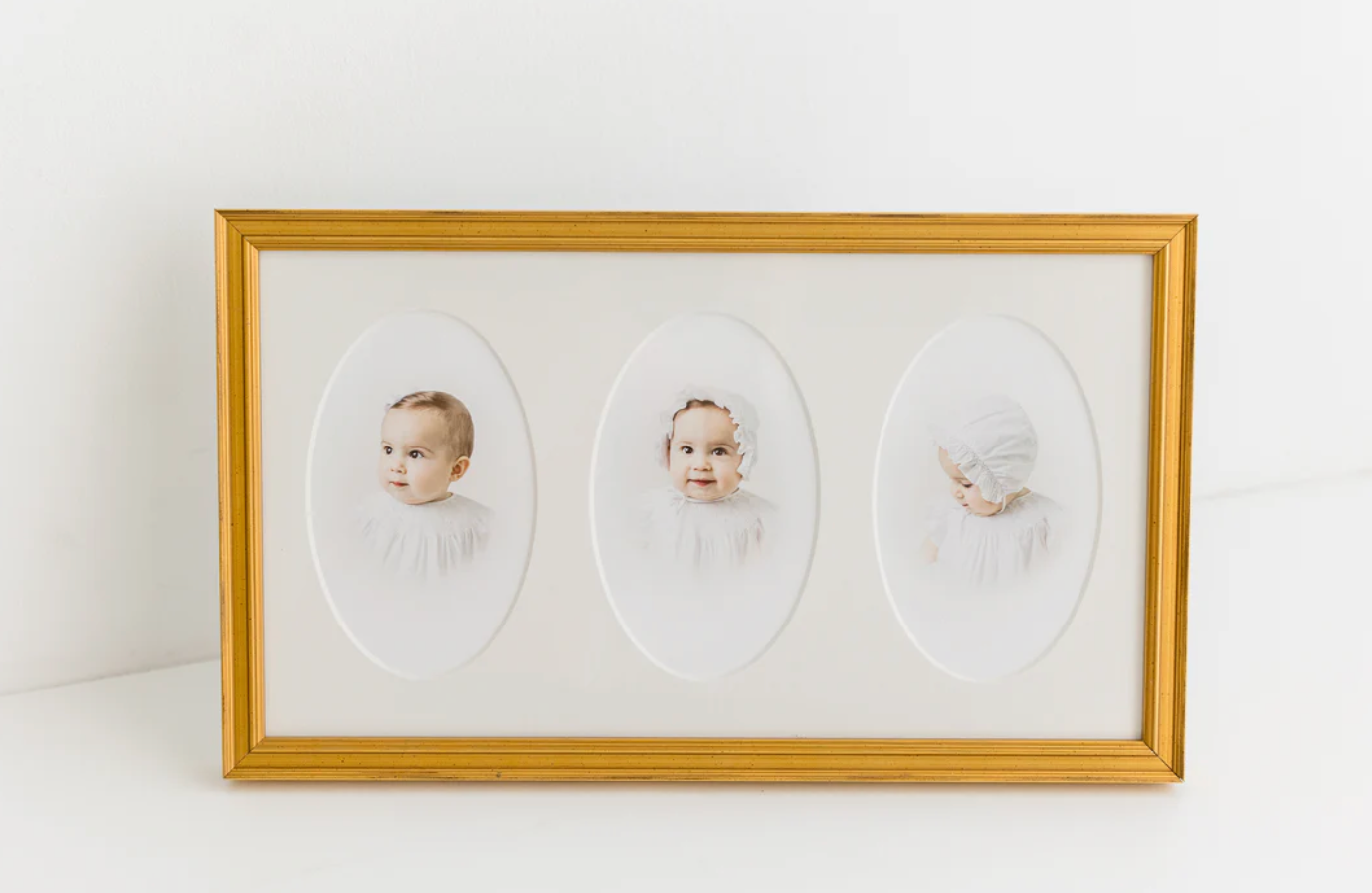 Gold framed triptych portrait of three babies in oval mats, displayed on white background.