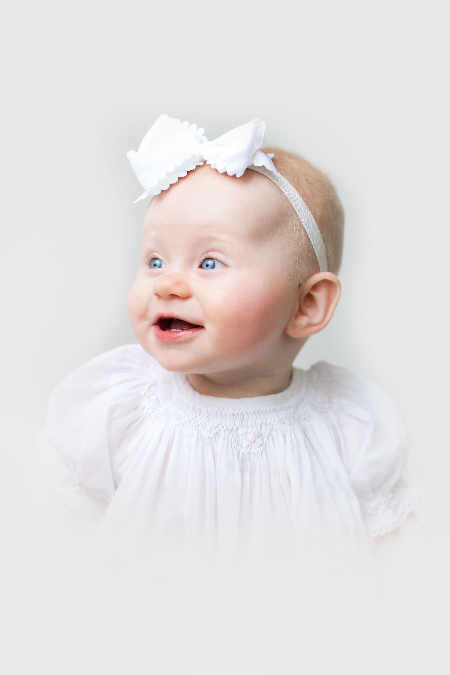 Smiling baby girl in white dress and bow headband with blue eyes against light background.