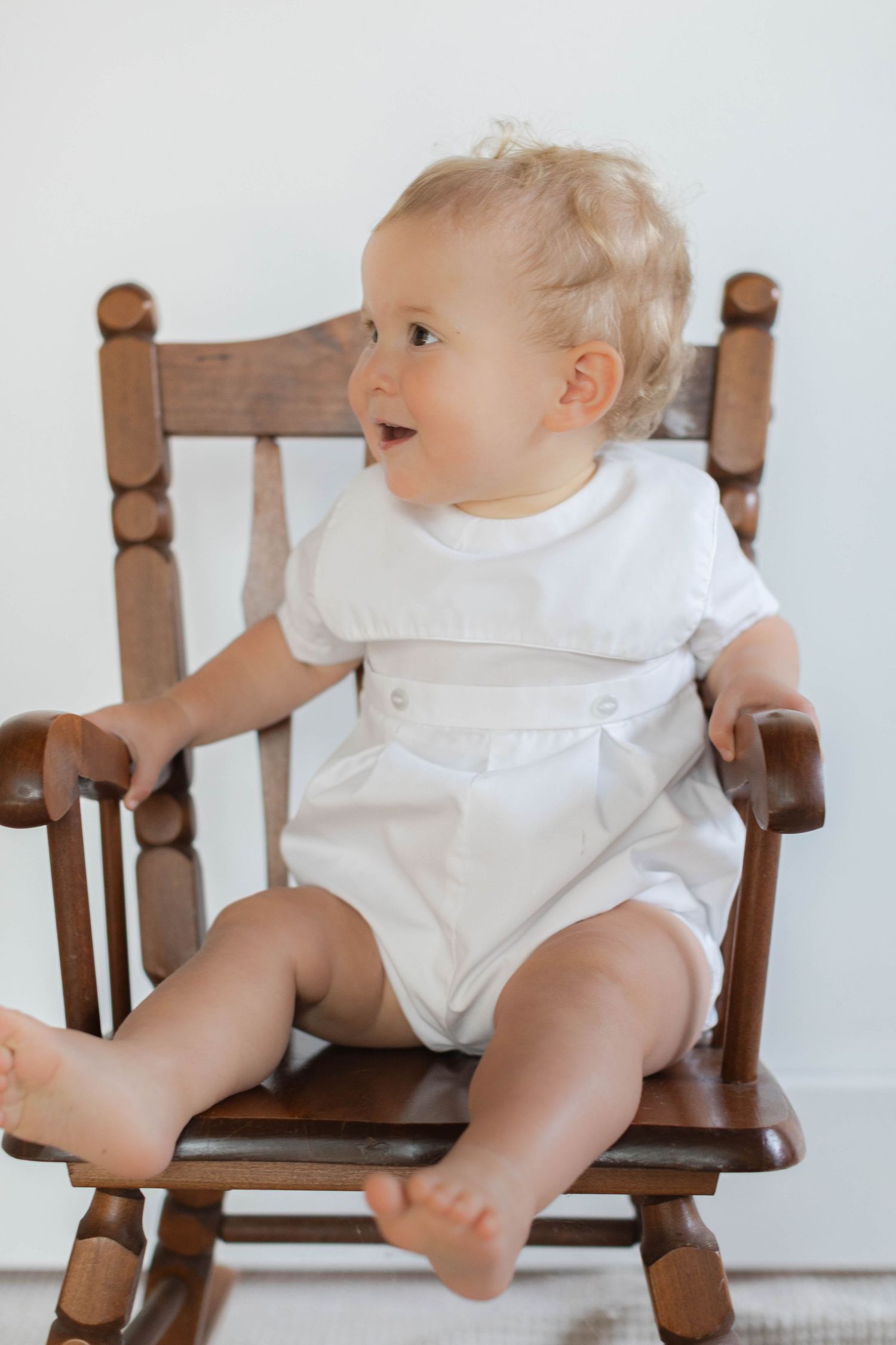 Baby in white romper sits in wooden rocking chair against white wall.