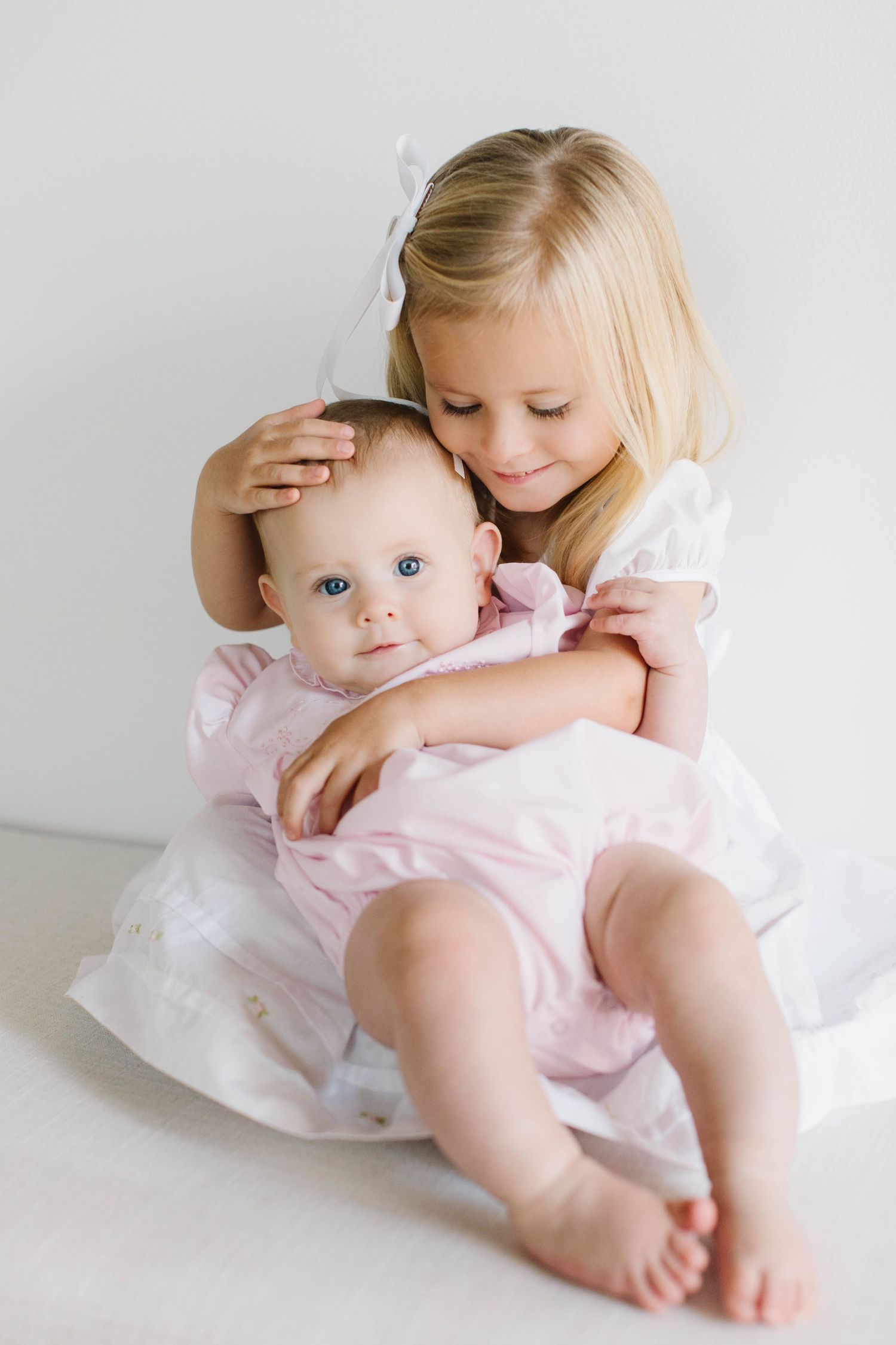 Two children embrace while wearing light colored clothing on a white backdrop.