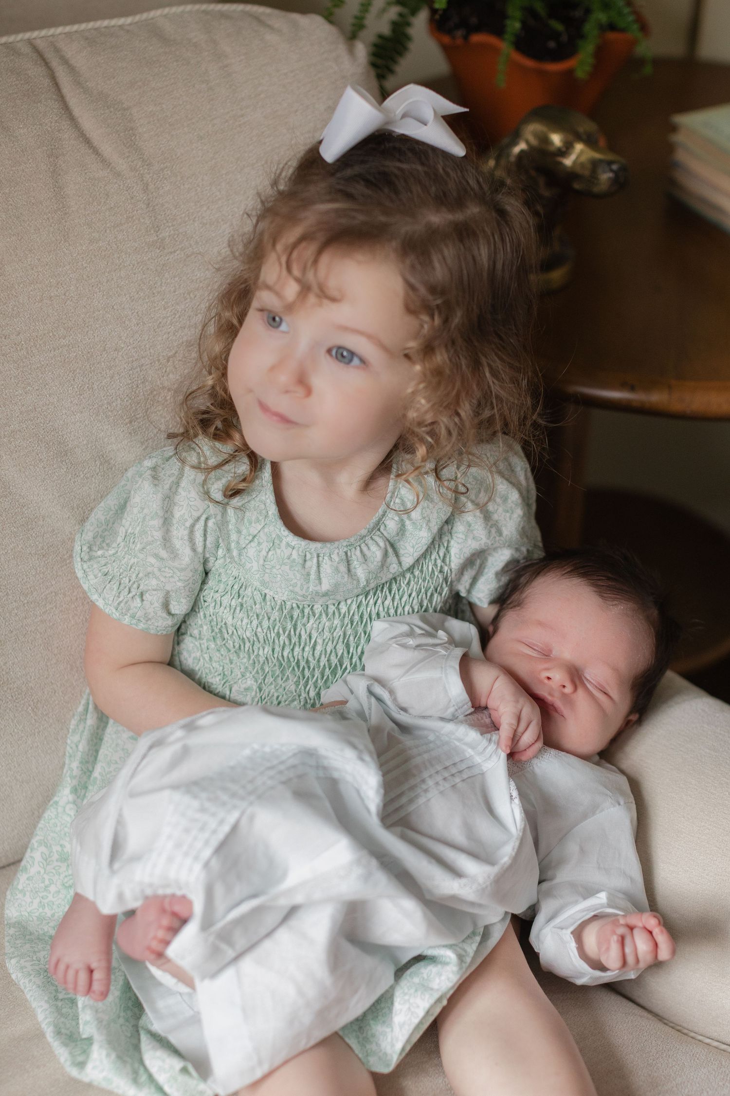Two children in light colored dresses relax together on a cream colored couch in a series of sweet candid moments.