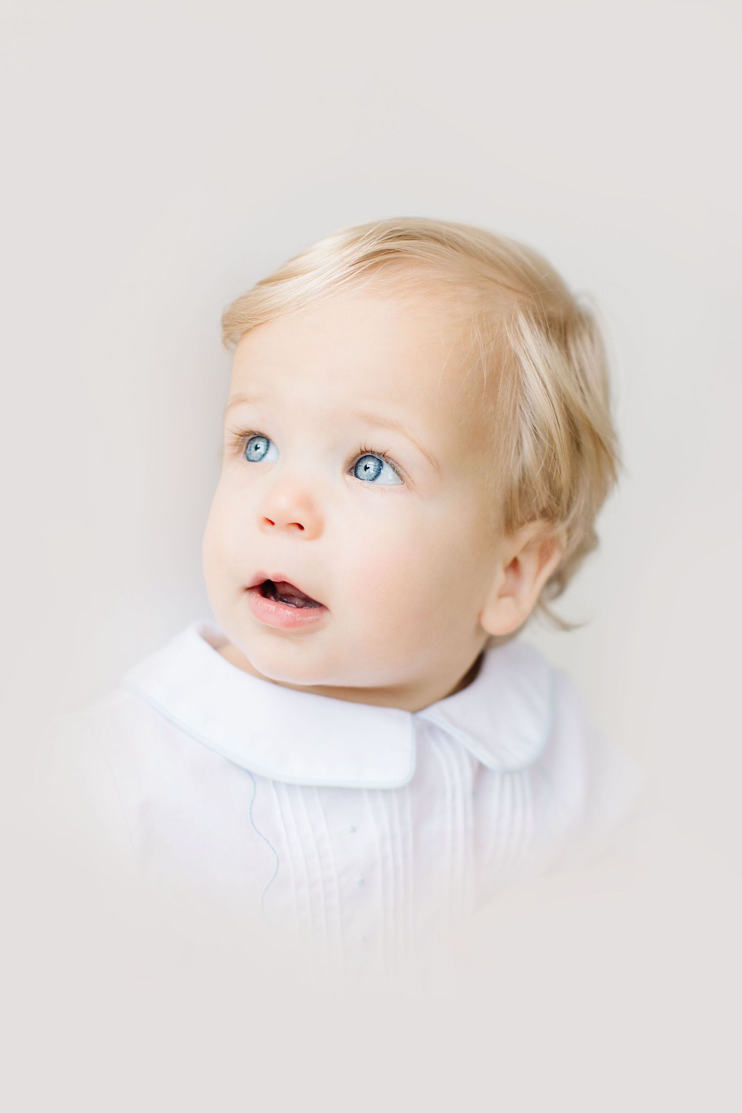 A series of three close-up portraits of a baby with blonde hair and blue eyes wearing white against a light background.