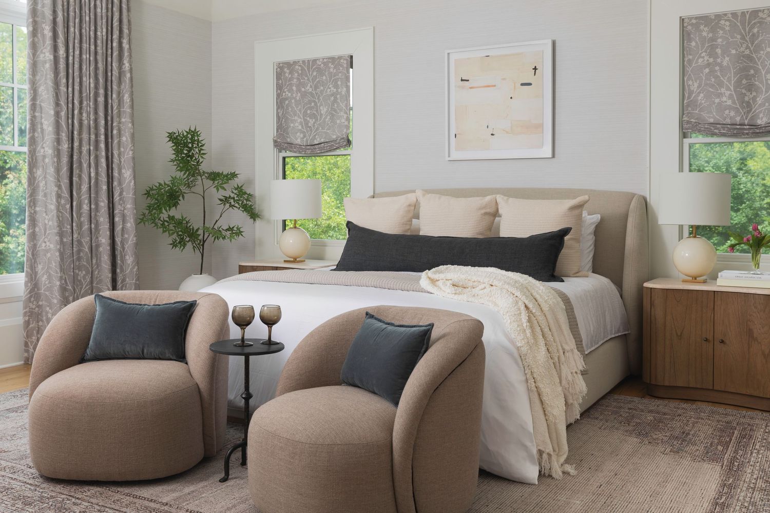 Modern bedroom with neutral tones, upholstered bed, curved accent chairs, and natural light from roman shade windows.