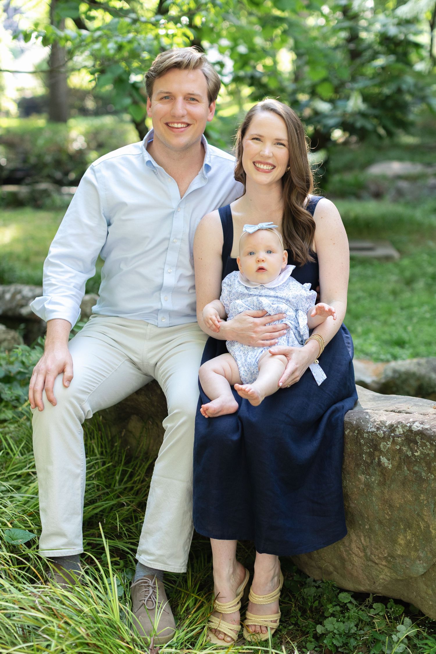 A smiling couple sits on a stone bench in a lush garden, holding their happy baby dressed in a white outfit.