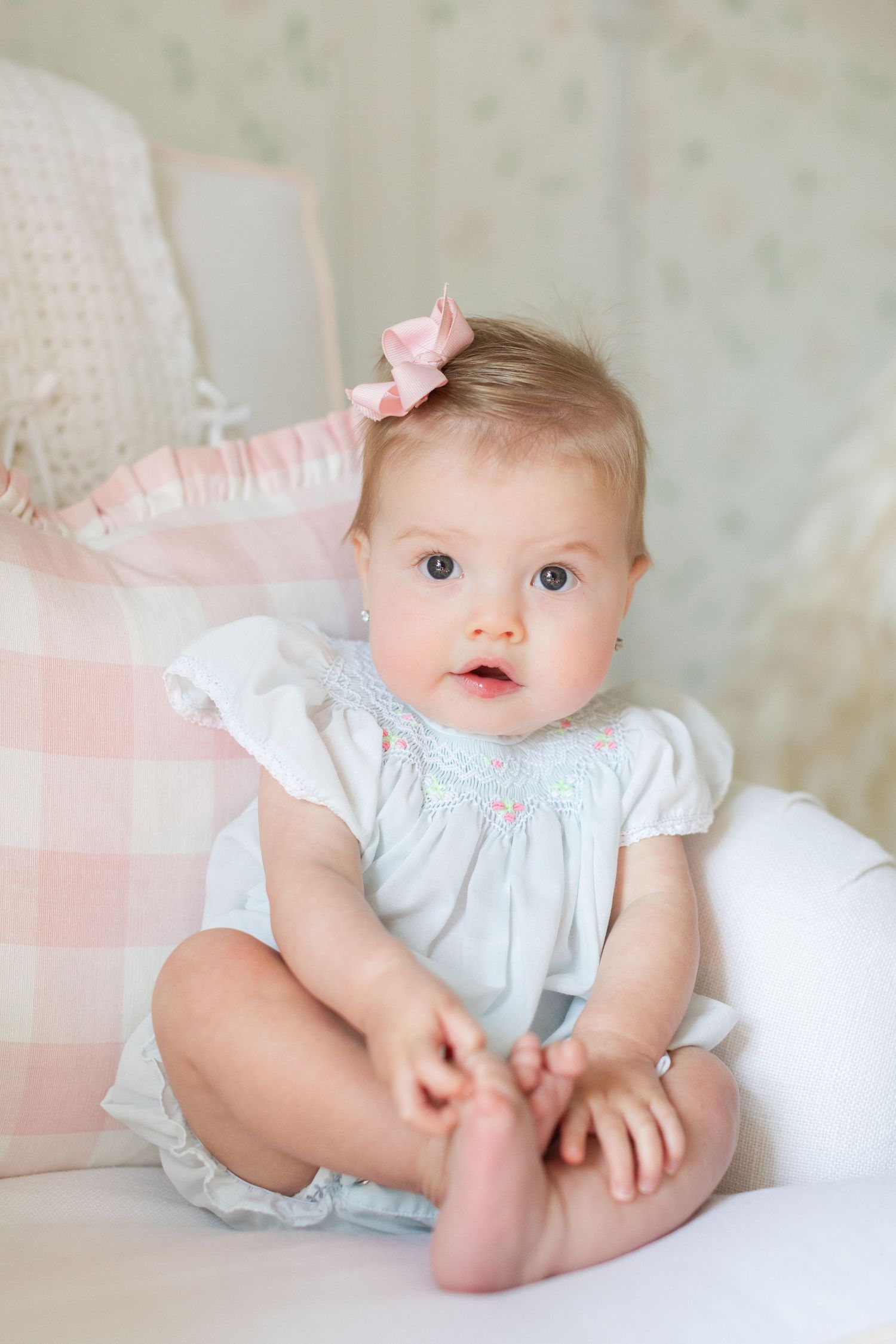 Adorable baby girl in white dress with pink bow sits on soft cushions, looking curiously at camera.