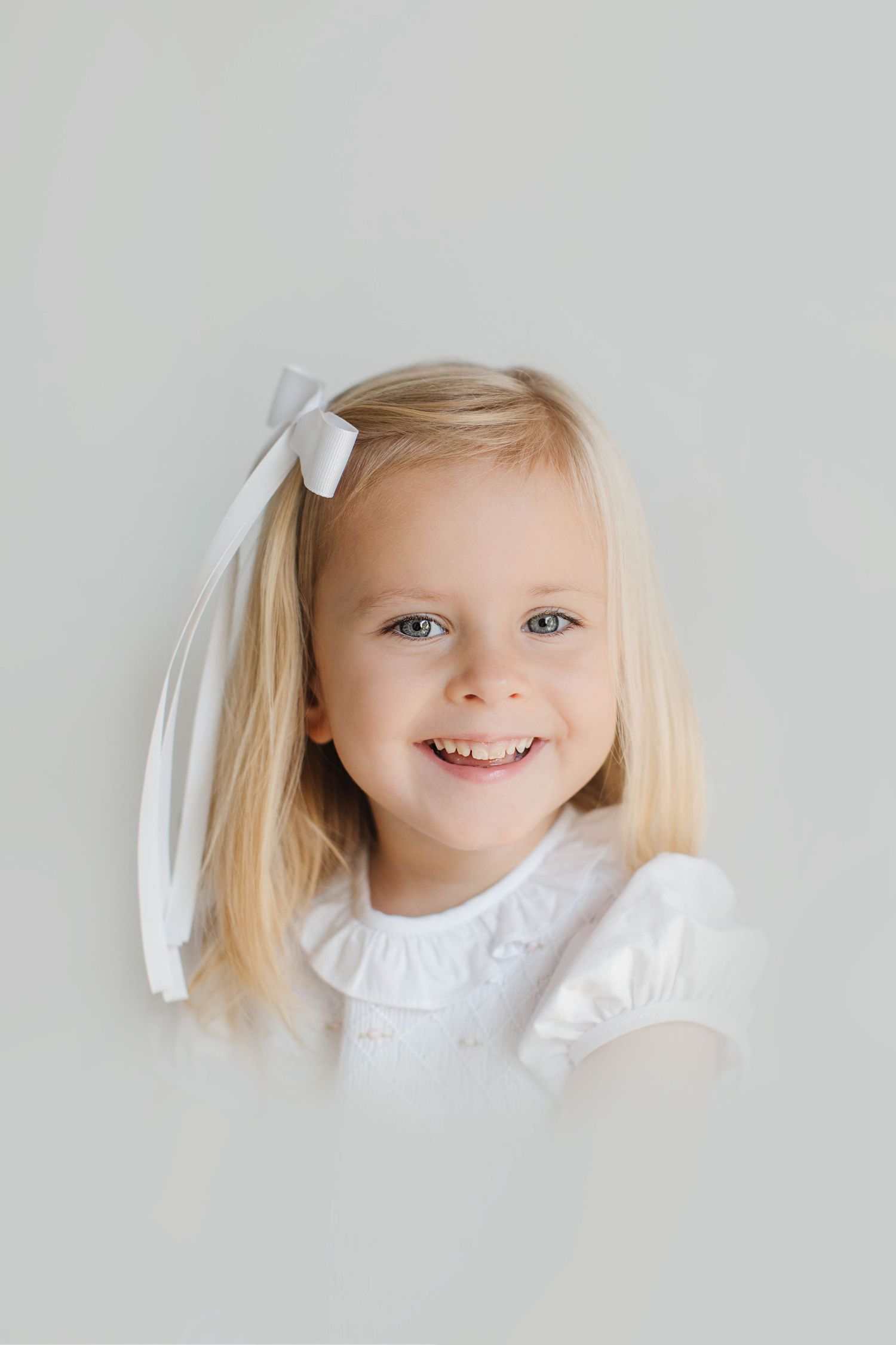 Heirloom portrait of a child with blonde hair and white bow smiling against a light background - Atlanta Heirloom Photography.