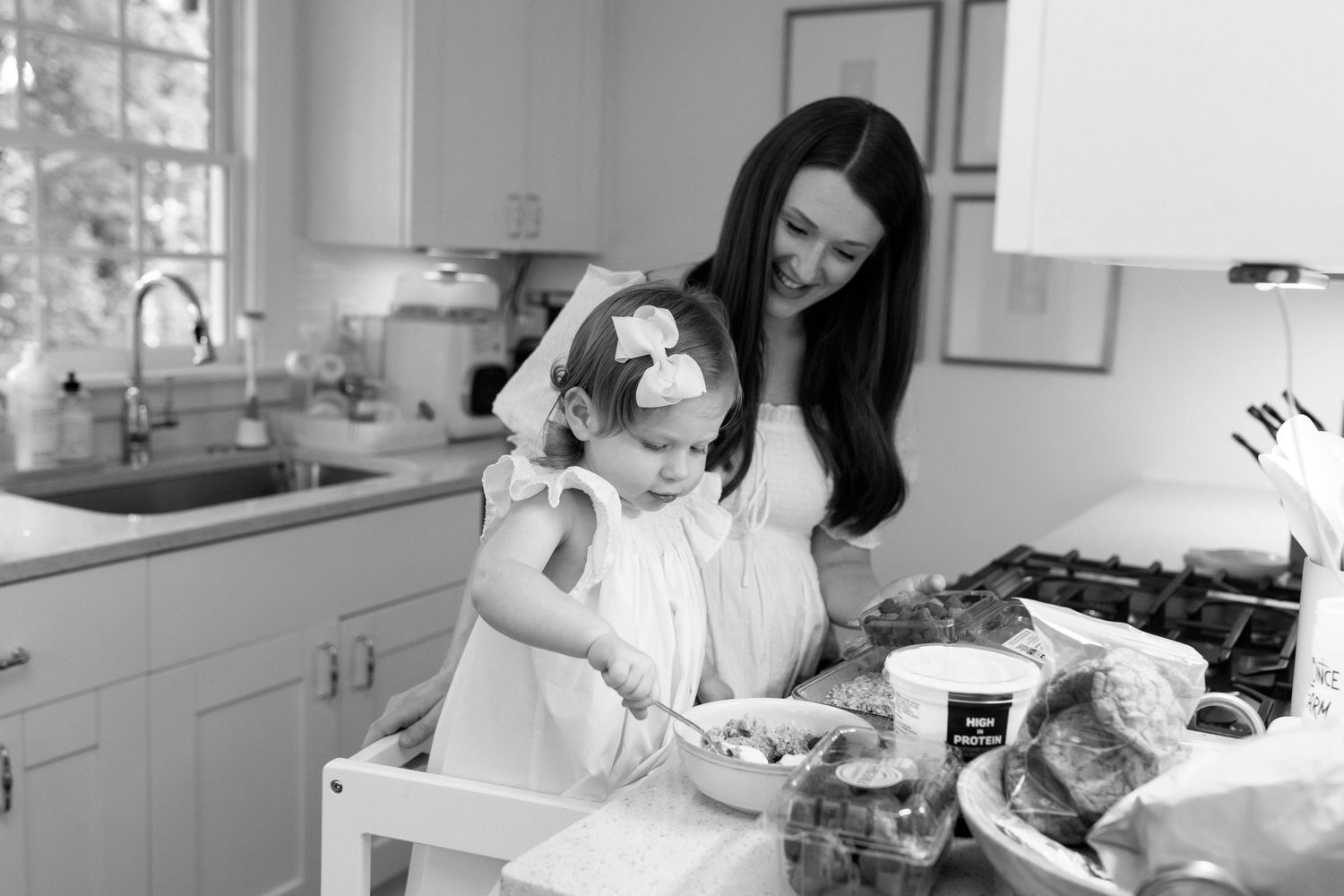 Parent and child in white dresses prepare food together in sunny kitchen.