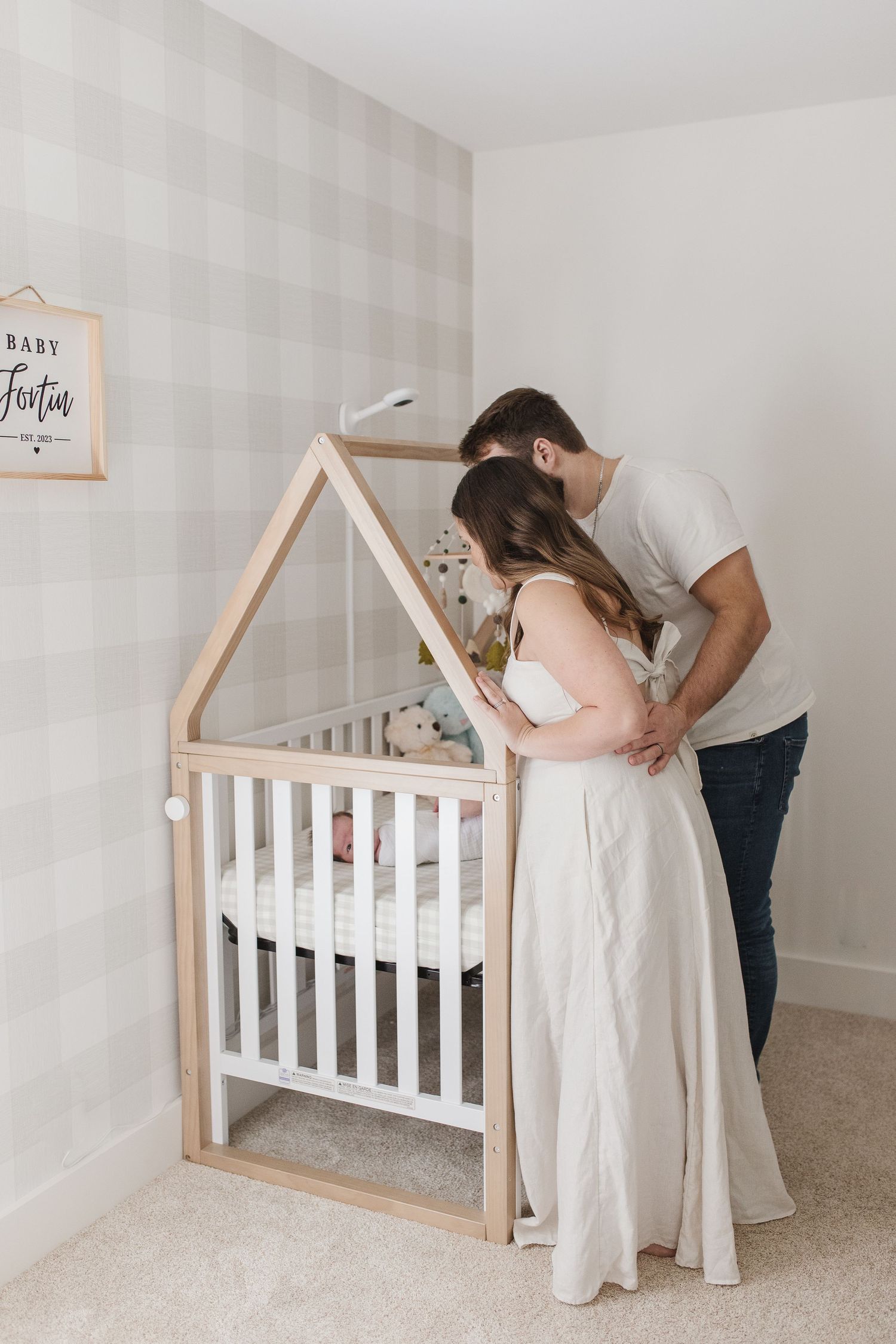 Parents placing baby in wooden house-frame crib in nursery with gray wallpaper.
