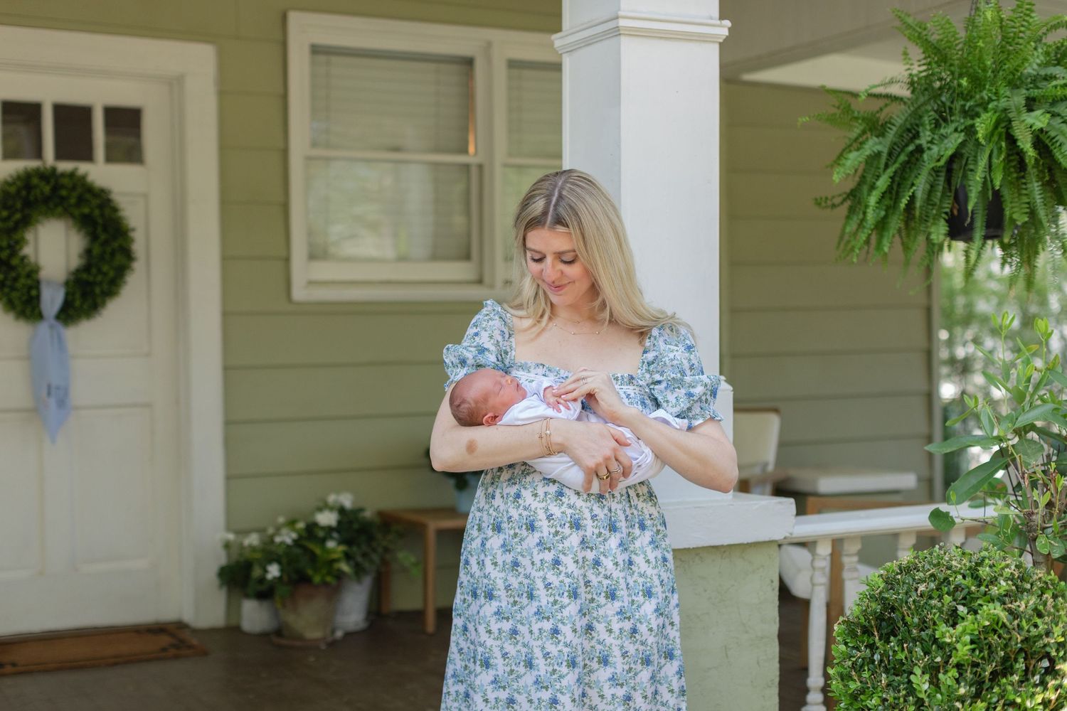 A mother in a floral dress holds her newborn baby on a charming green home porch with lush plants.