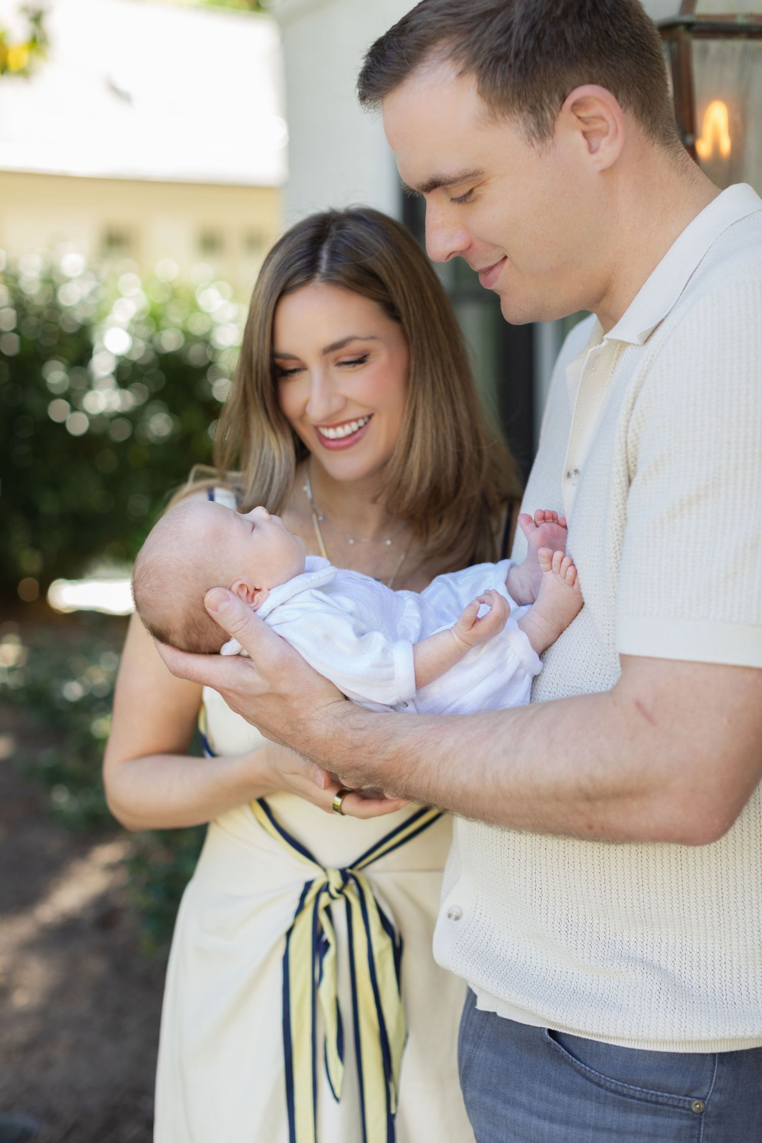 Smiling parents lovingly hold newborn baby outdoors in front of home surrounded by greenery.