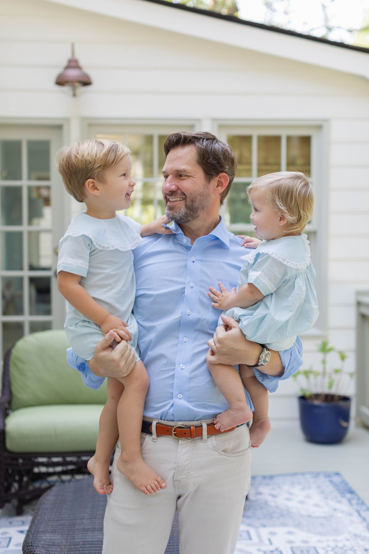 A smiling parent in blue dress shirt holding twin toddlers in coordinating light blue outfits on a deck.