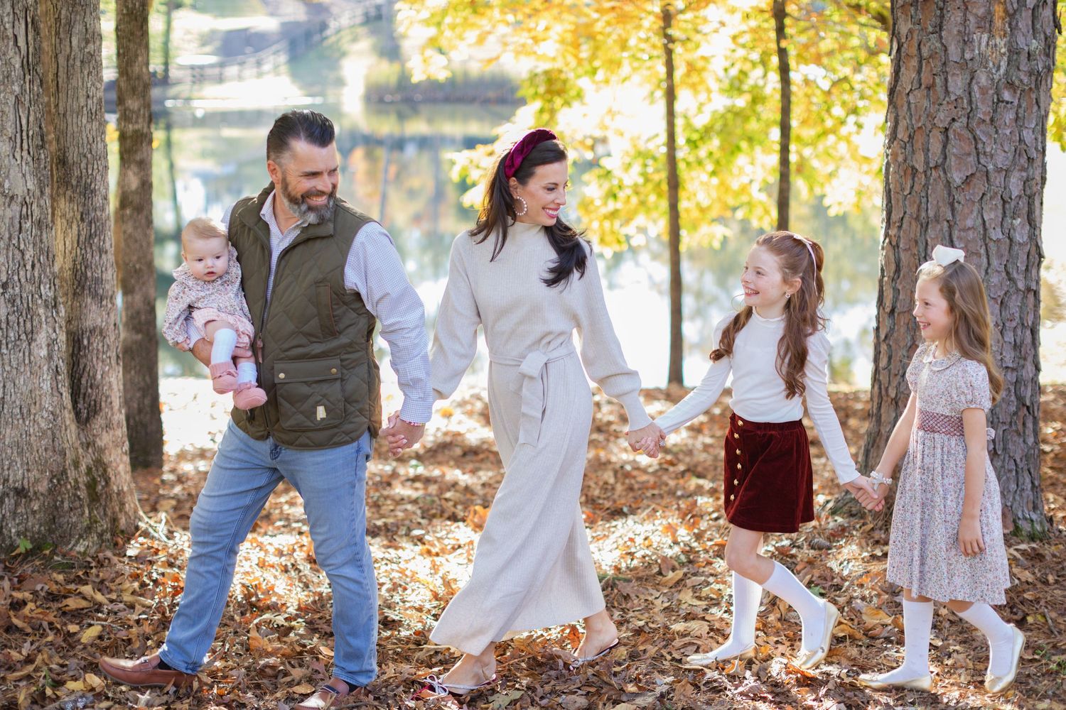 A family in coordinating autumn outfits walks together through a forest path with golden fall foliage.