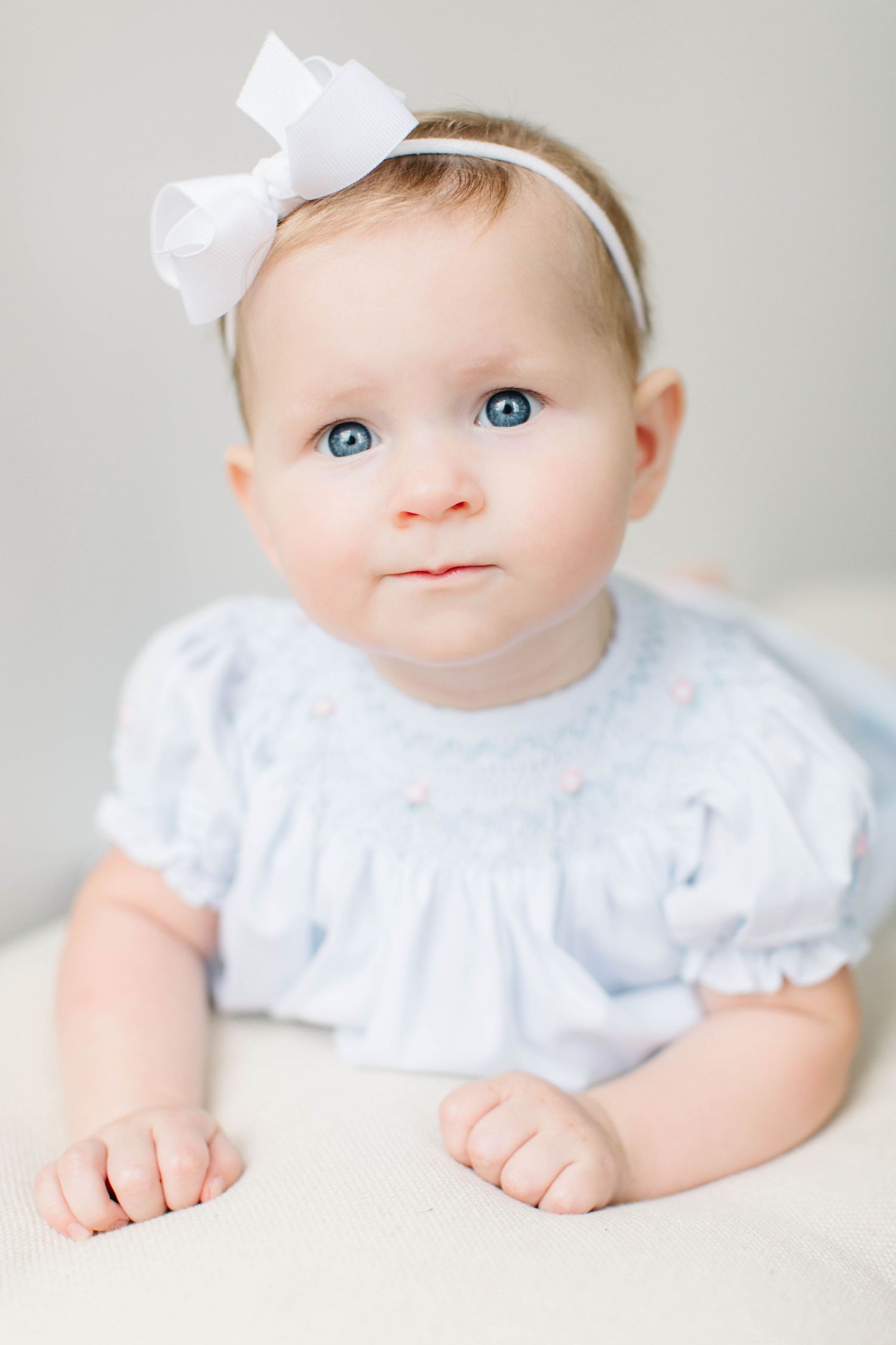 A baby in white lace dress with bright blue eyes and a white bow headband rests on a white background.