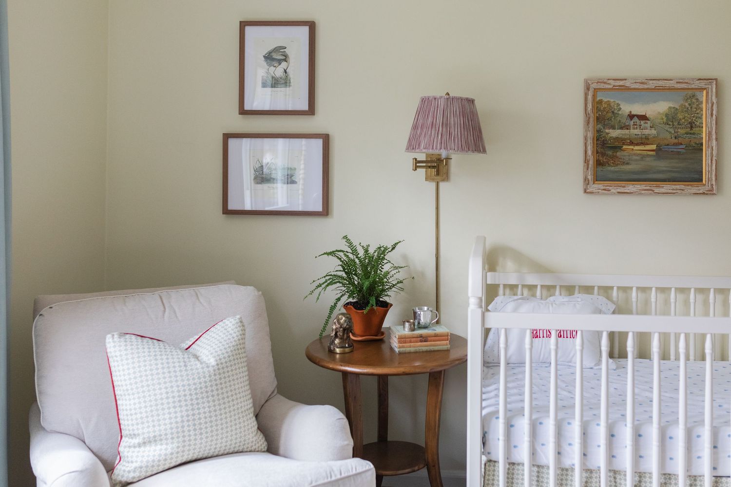 A cozy nursery room with white crib, framed artwork, table lamp, and potted plant against neutral walls.