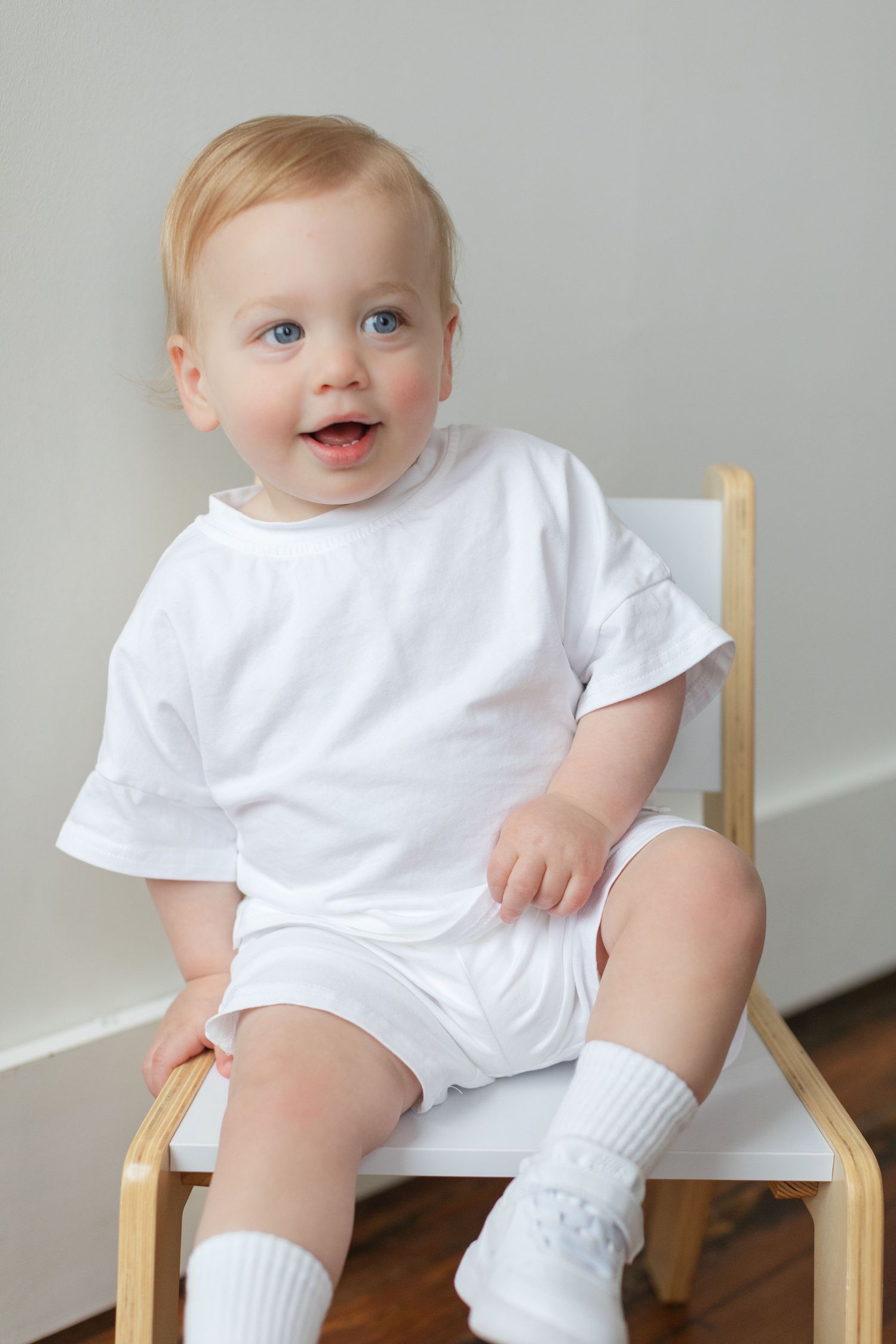 Happy baby wearing all white outfit and socks sits on wooden chair against light gray wall.