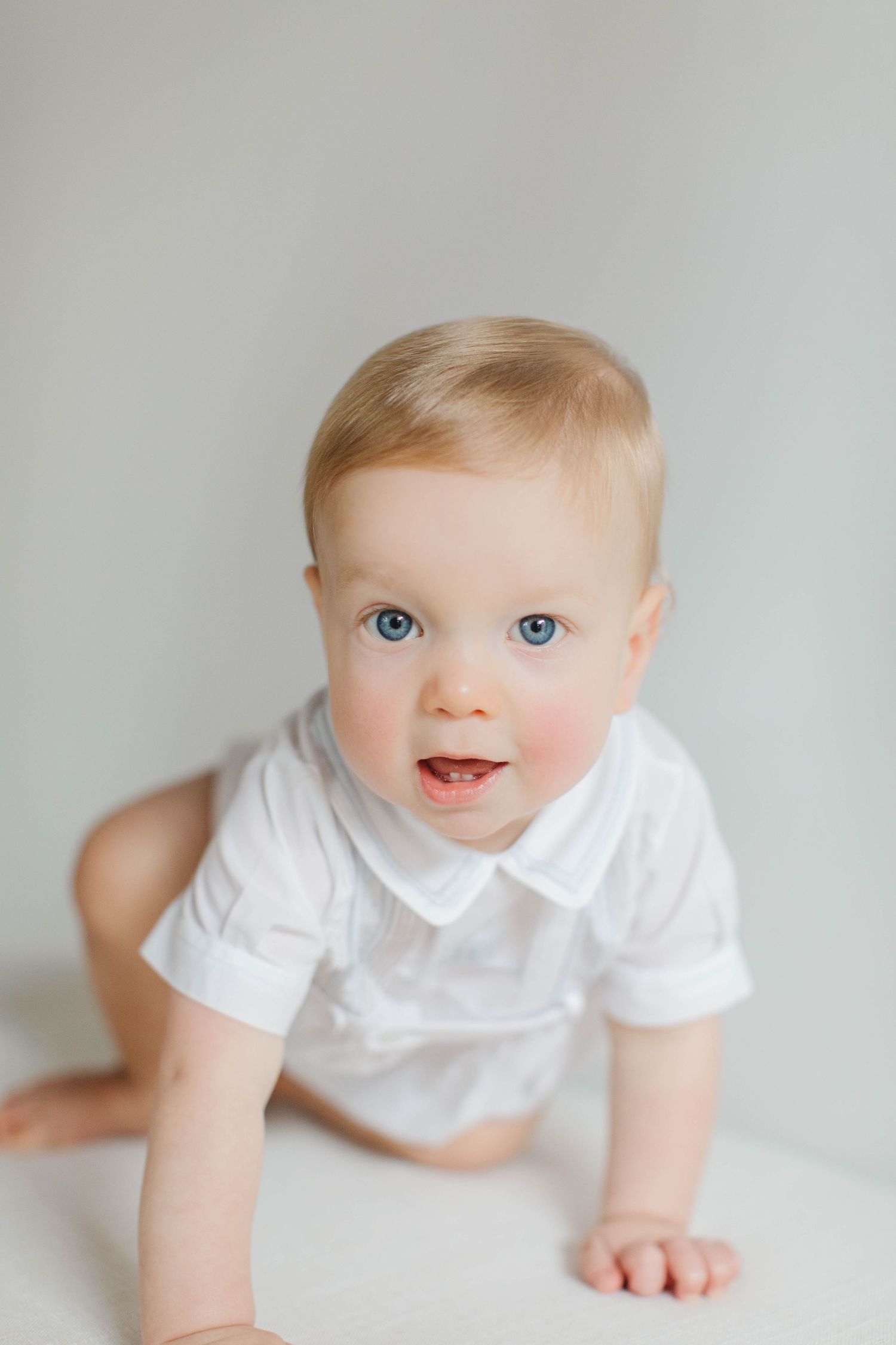 Baby in white collared shirt crawling on white surface with bright blue eyes and joyful expression.