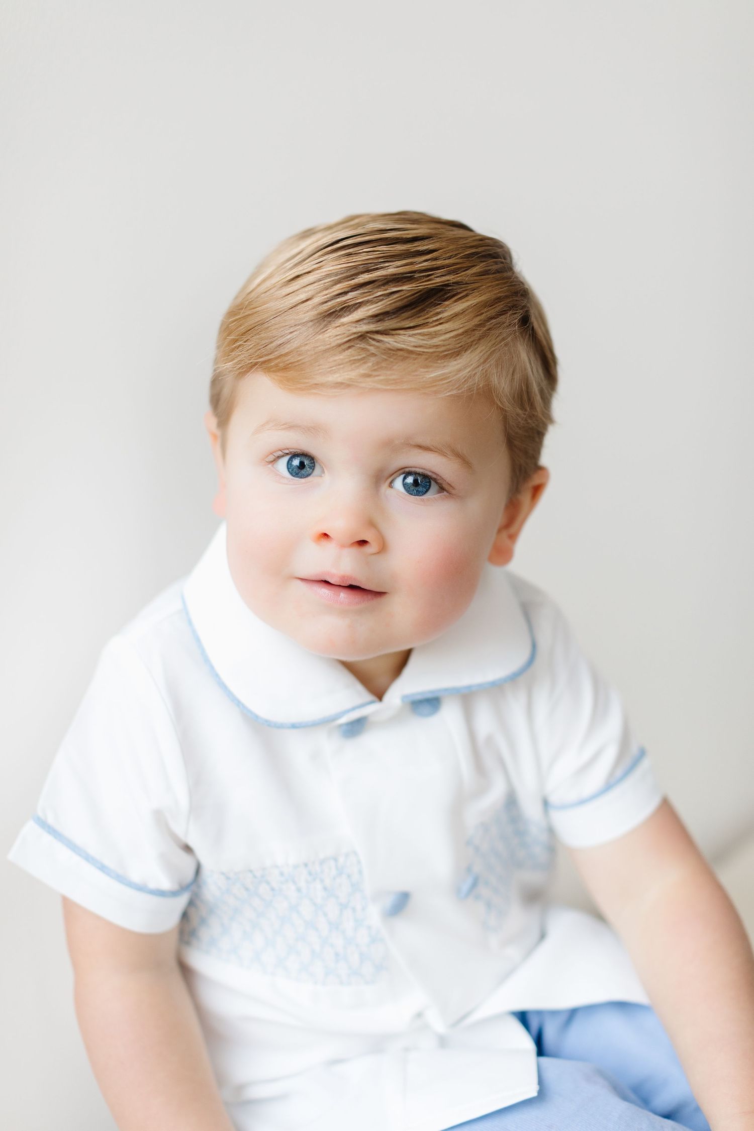 A baby in a white collared shirt with blue trim smiles sweetly while lying on a light background.