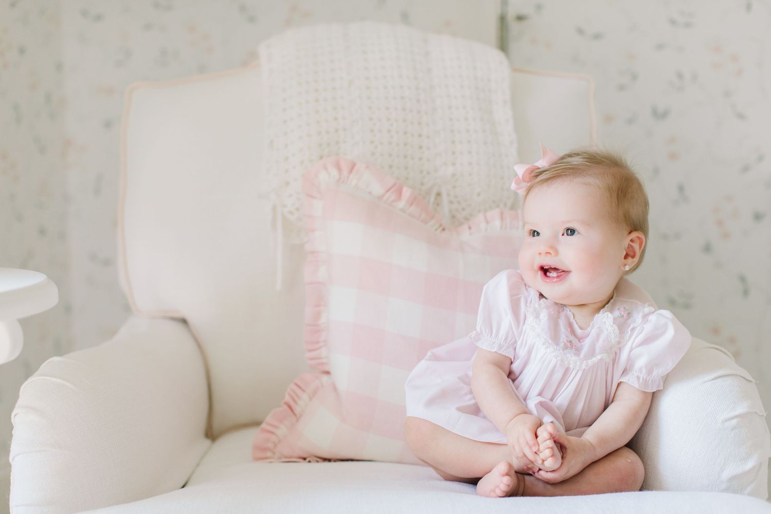 Smiling baby girl in pink outfit sitting on white chair with pink gingham pillow in soft nursery setting.