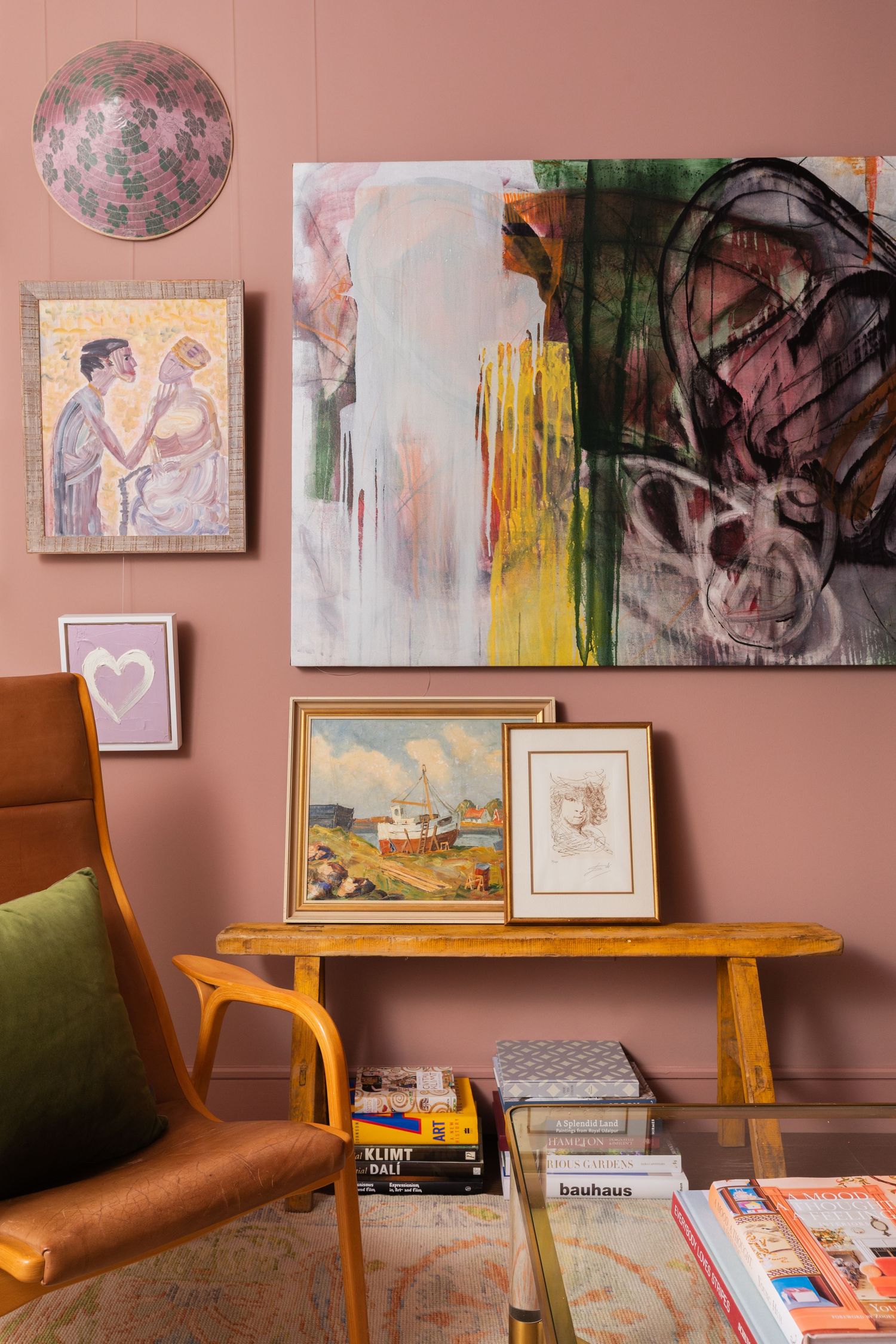 Eclectic living room with pink walls, abstract art, framed paintings on a wooden bench, and a mid-century modern orange armchair.