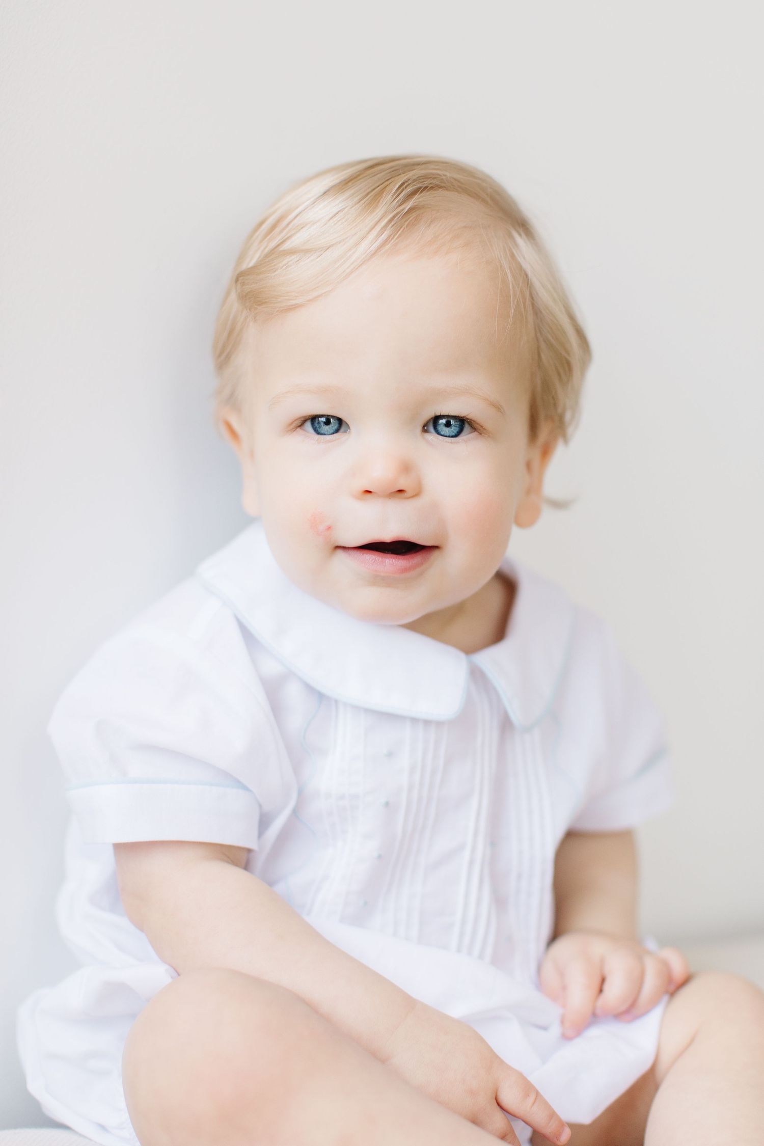 Baby in white collared romper smiles while sitting against light background.
