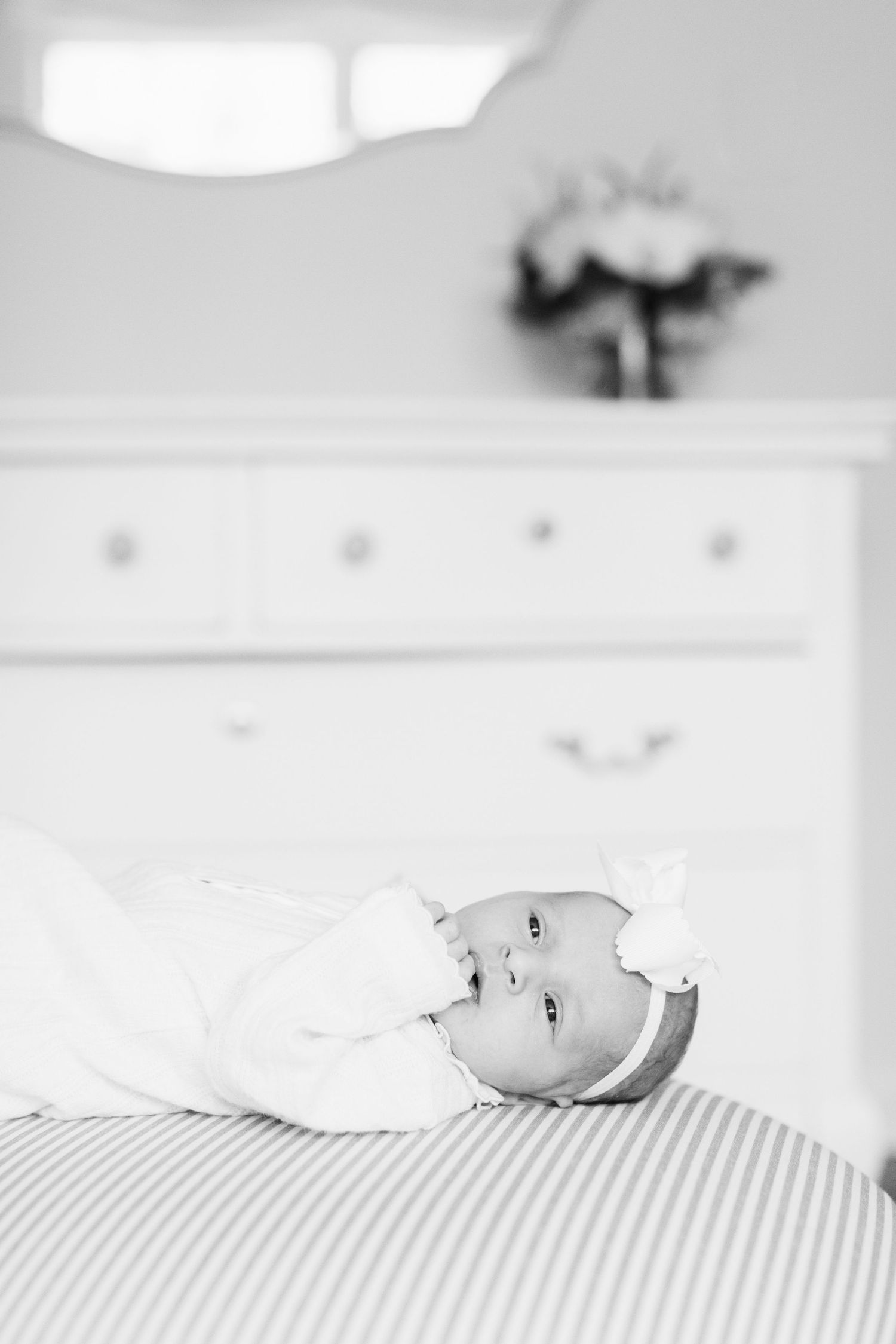Newborn baby in white outfit lying on striped bedding with white dresser in background.