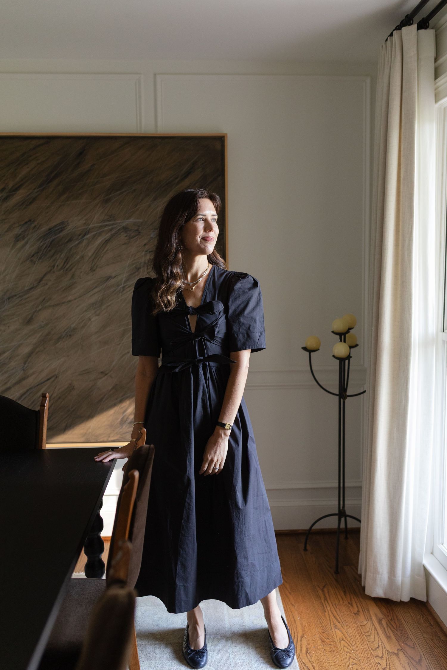 Woman in elegant black midi dress with bow detail and puff sleeves standing in a sunlit room.